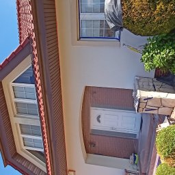 Firma remontowo-wykończeniowa - Elewacja domu w trakcie malowania: okna zabezpieczone folią, widoczne narzędzia malarskie, świeżo pomalowana ściana. Częściowo zakryte krzewy.