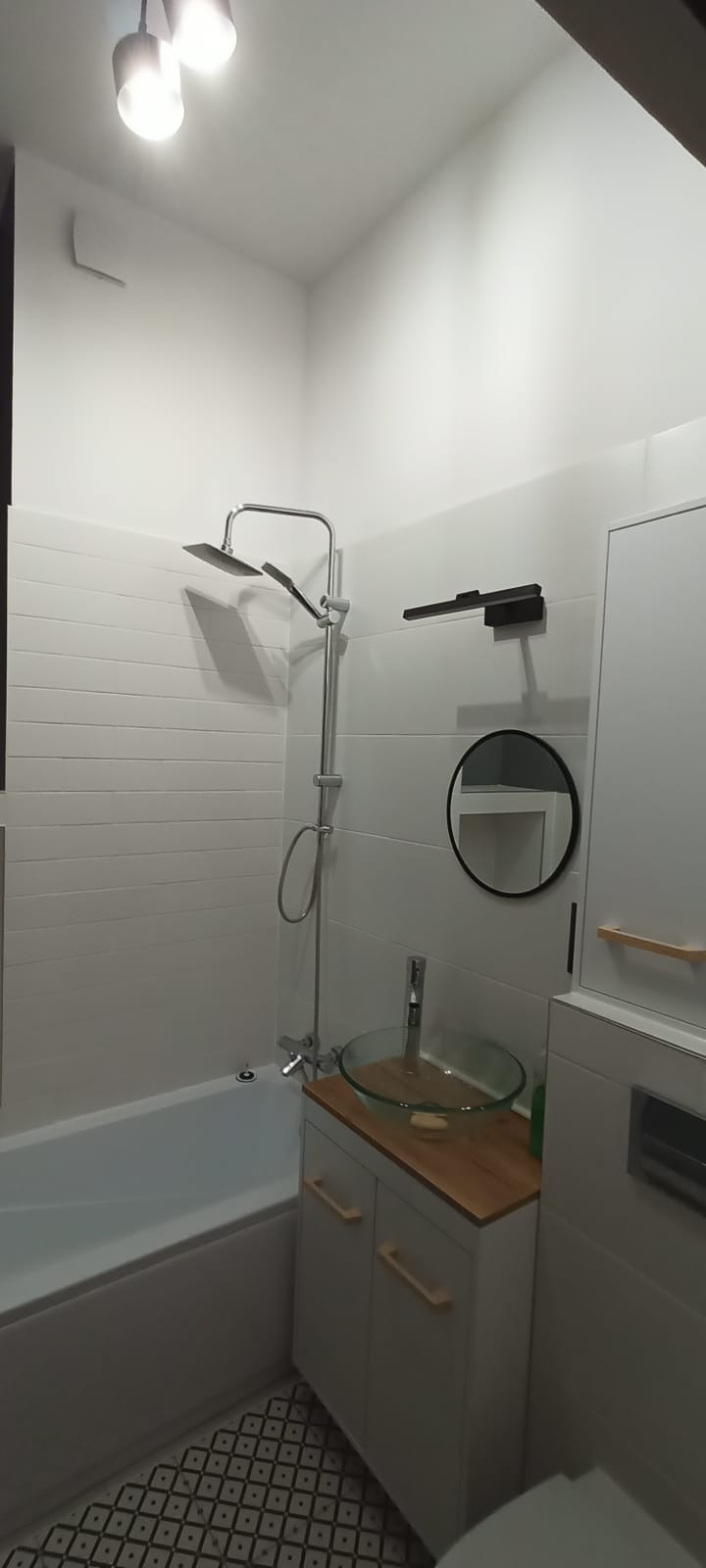 Nowoczesna łazienka z białą armaturą, szklaną umywalką na drewnianym blacie, baterią chromowaną, kabiną prysznicową z deszczownicą i wzorzystą podłogą.