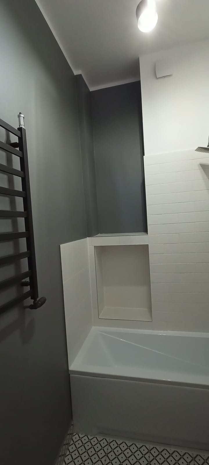 Wykończona łazienka z wanną, zabudowaną wnęką z półkami, grzejnikiem drabinkowym w kolorze antracytowym i płytkami imitującymi drewno na ścianie.