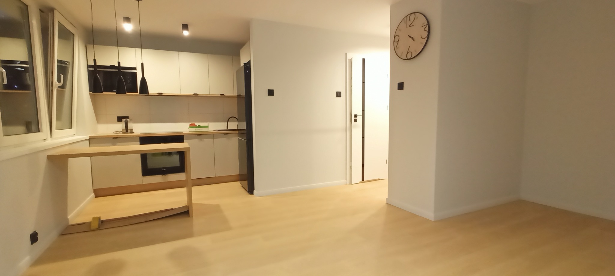 Jasne, przestronne wnętrze mieszkania z minimalistyczną kuchnią z białymi szafkami i drewnianymi blatami, widoczne okno, lampy wiszące, duży zegar na ścianie i drewniana podłoga.