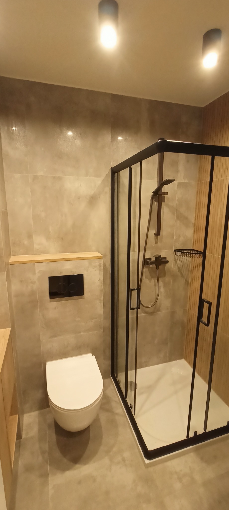 Nowoczesna łazienka z kabiną prysznicową w czarnej ramie, podwieszaną toaletą i płytkami imitującymi beton, oświetlona ciepłym światłem.