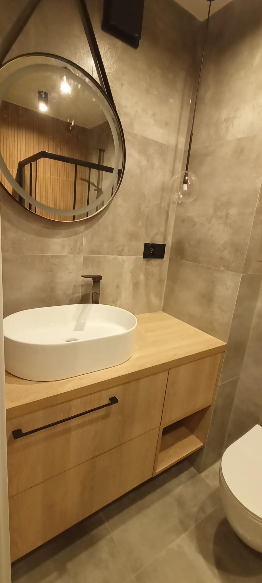 Nowoczesna łazienka z okrągłym lustrem z podświetleniem, wiszącą szafką z drewnianym blatem, ceramiczną umywalką nablatową i minimalistyczną armaturą w kolorze czarnym, uzupełniona szklaną lampą...