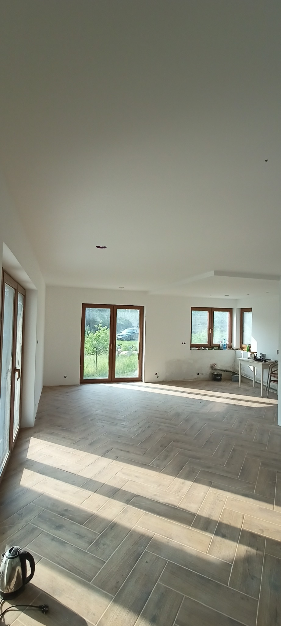 Jasne wnętrze po remoncie z parkietem w jodełkę, duże okna z widokiem na zieleń. Widoczny czajnik elektryczny. Świeżo pomalowane ściany na biało.