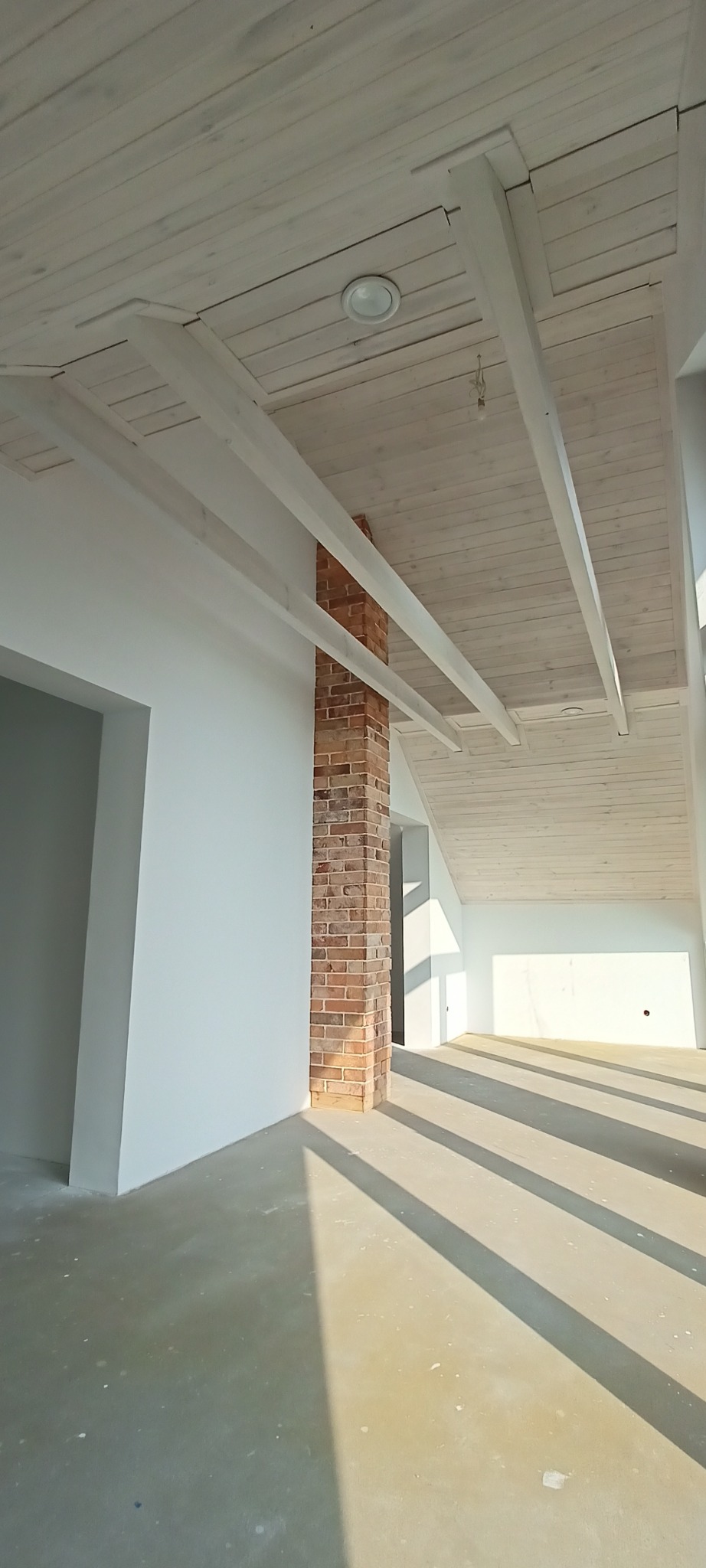Jasne wnętrze z ceglanym kominem i drewnianym sufitem. Widoczne białe ściany, betonowa podłoga i promienie słońca wpadające przez okno. Surowy stan wykończenia.