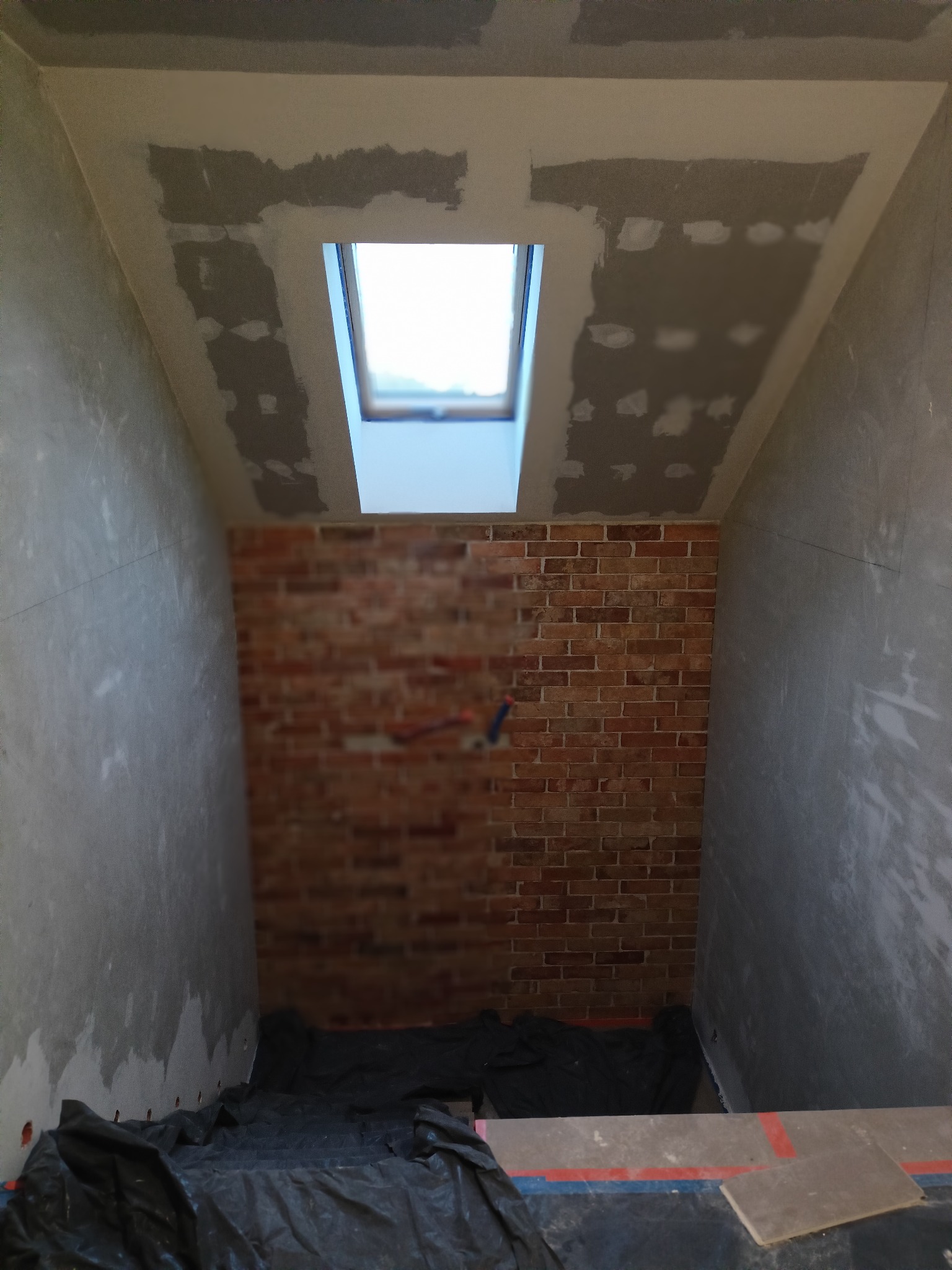 Poddasze w trakcie prac wykończeniowych: ściany przygotowane do malowania, fragment ściany z cegły, okno dachowe. Widoczne ślady szpachlowania.