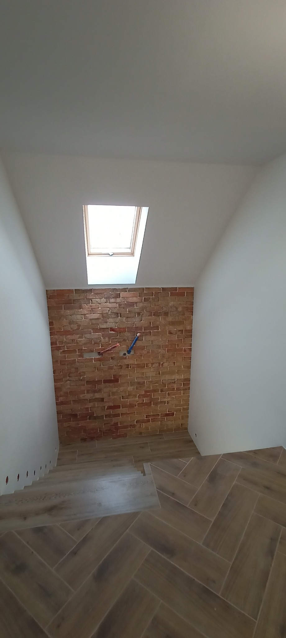 Klatka schodowa z ceglaną ścianą i drewnopodobnymi płytkami ułożonymi w jodełkę. Widoczne okno dachowe i wystające rury instalacji. Nowoczesne wykończenie.