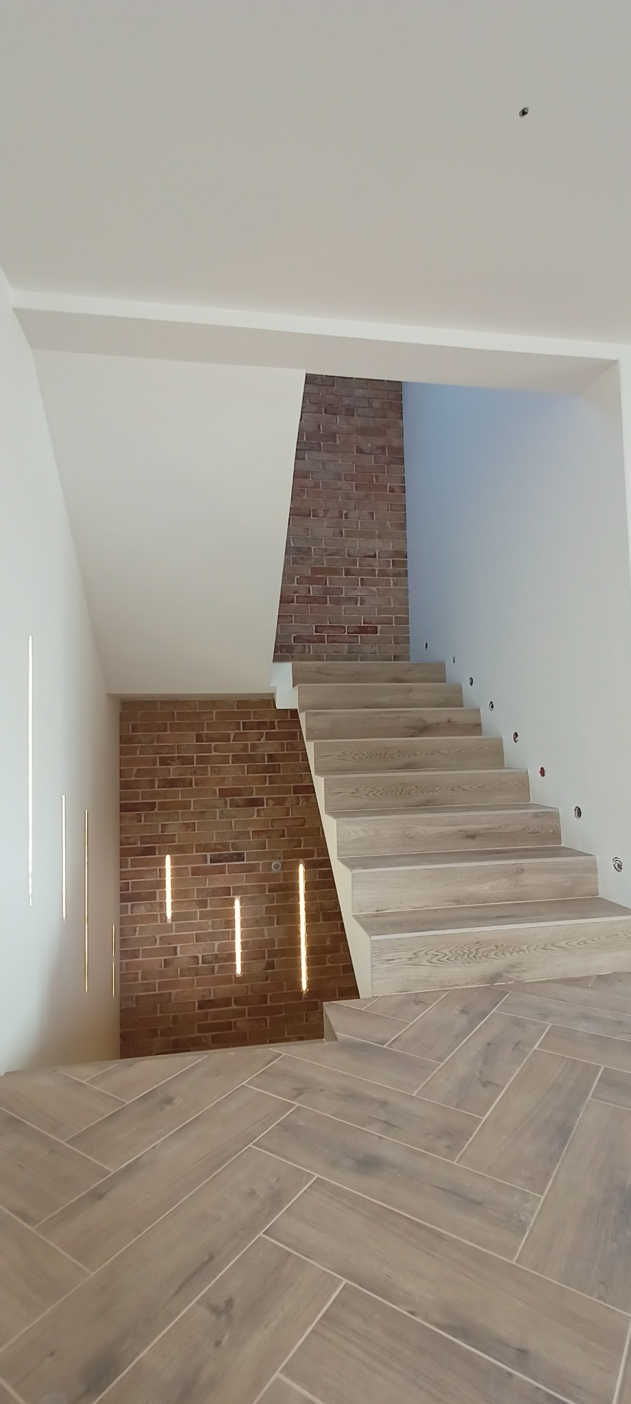 Schody z drewnianymi stopniami i ceglaną ścianą z oświetleniem punktowym. Podłoga wyłożona płytkami imitującymi drewno w jodełkę. Nowoczesne wykończenie wnętrza.
