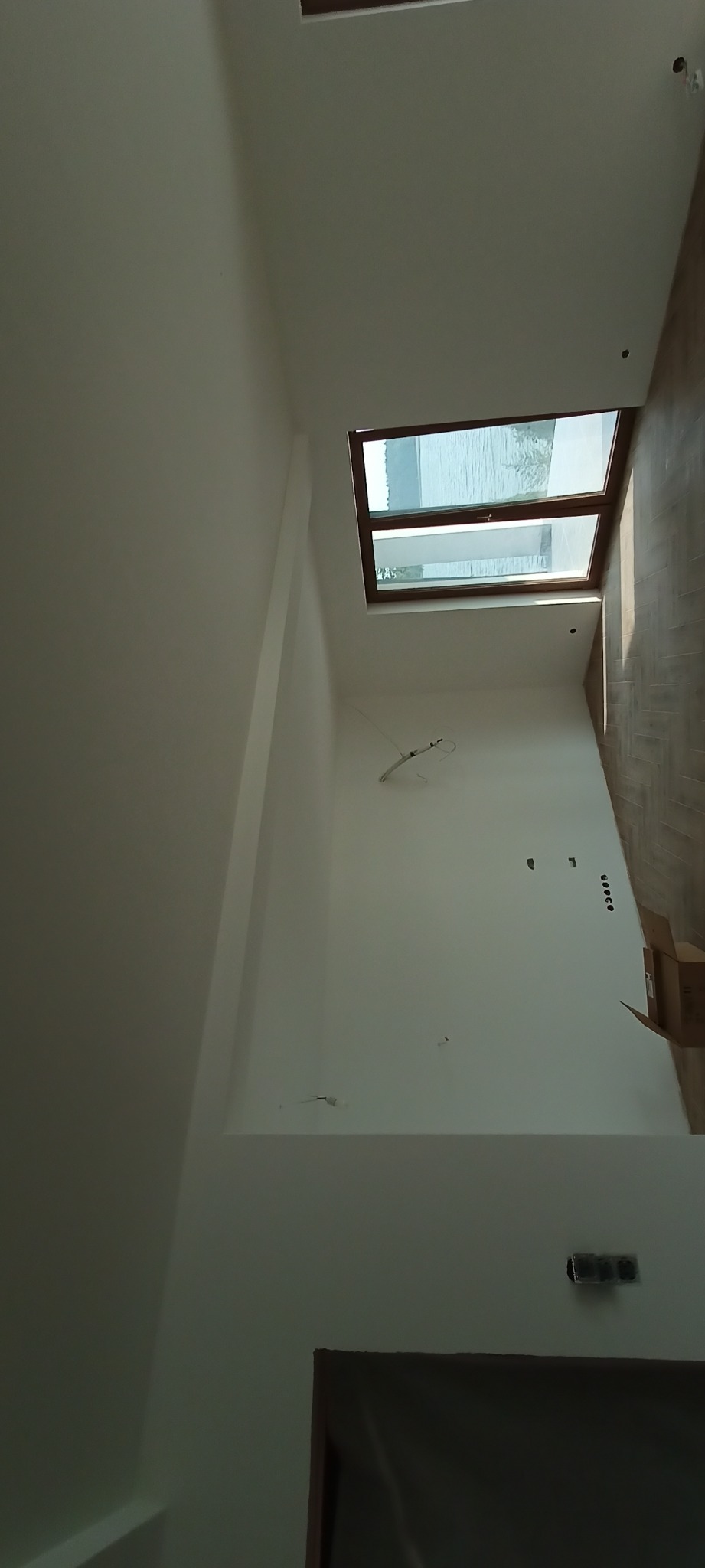 Wnętrze w trakcie remontu: białe ściany, okno dachowe z widokiem na zewnątrz, fragment podłogi z paneli, widoczne puszki elektryczne i kable.