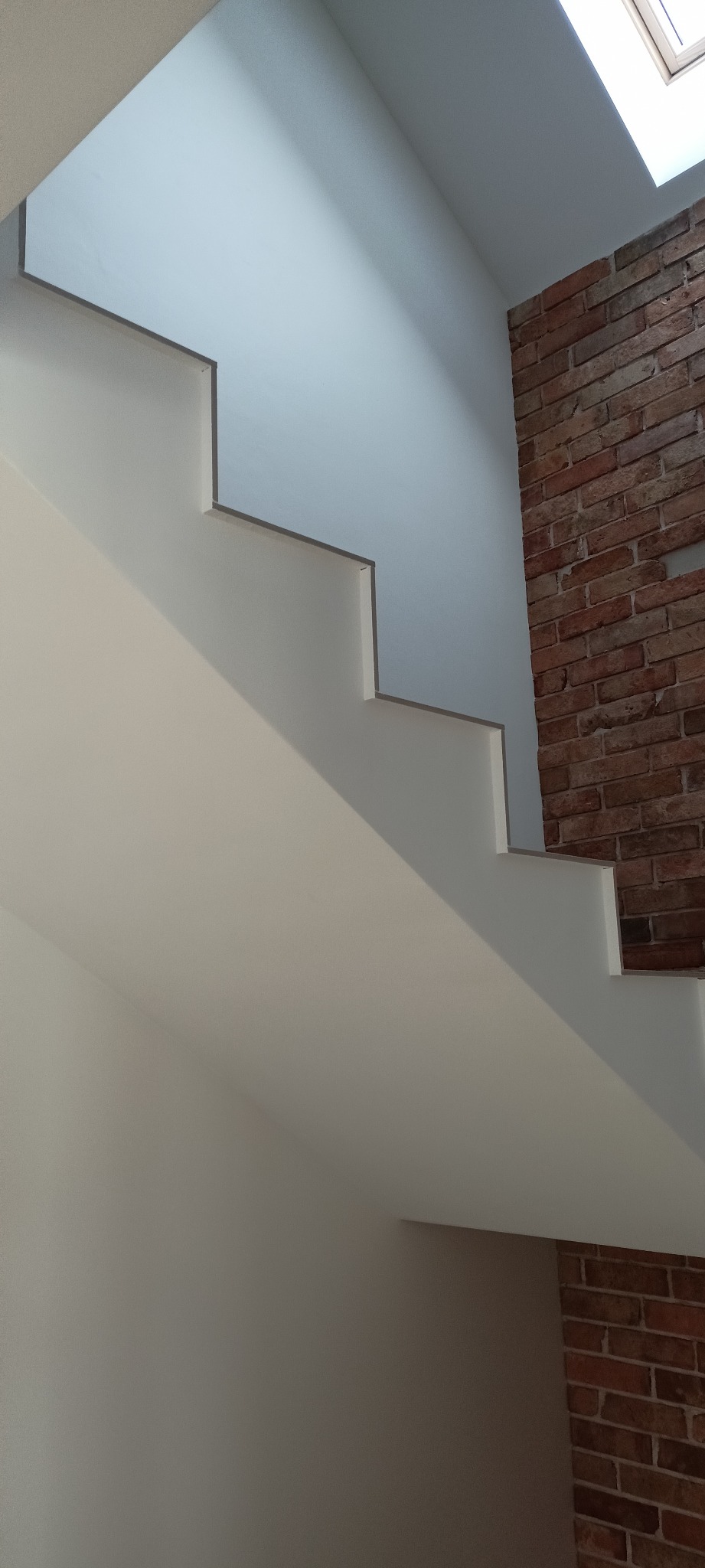 Białe schody z minimalistycznym wykończeniem, kontrastujące z fragmentem ściany z czerwonej cegły. U góry widoczne okno dachowe wpadające do wnętrza.