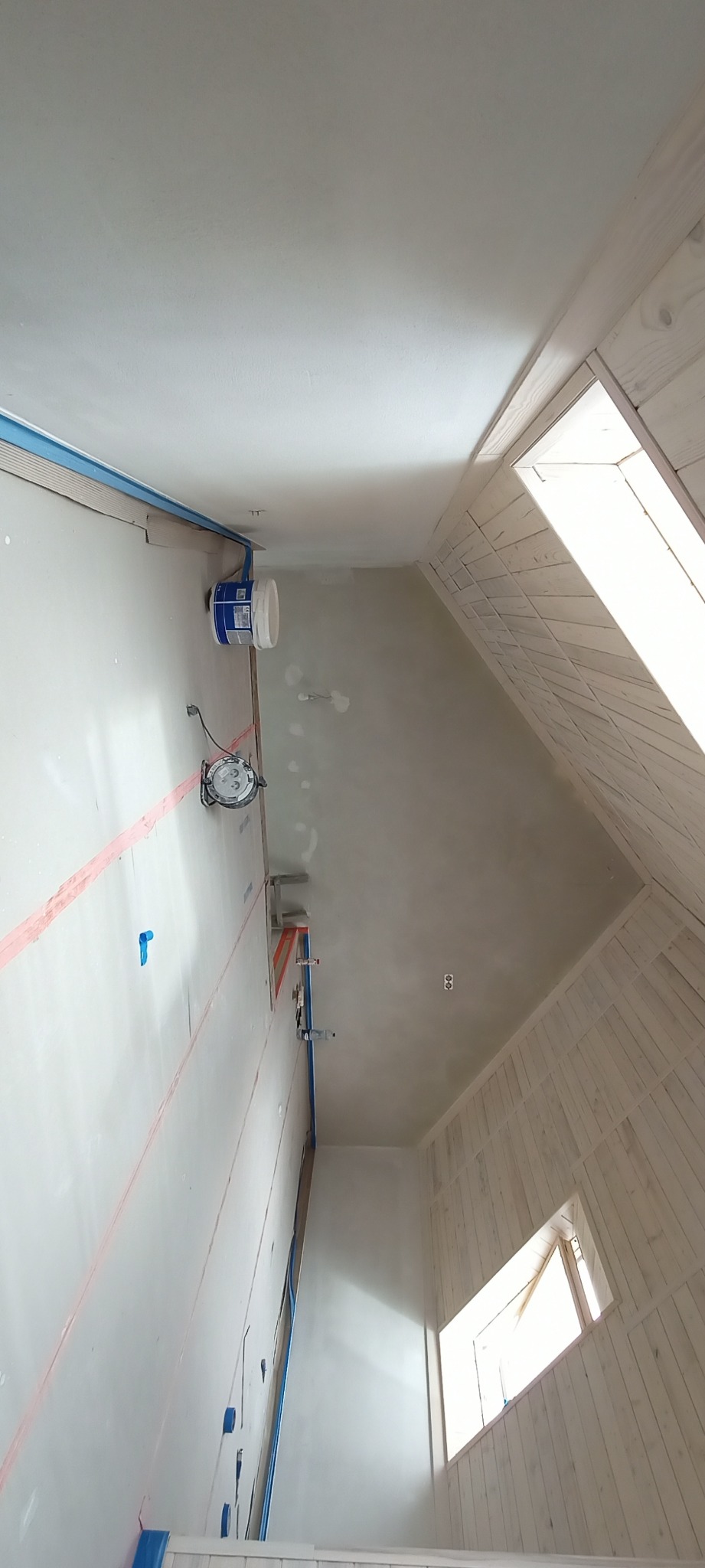 Poddasze w trakcie wykańczania: ściany z płyt gipsowych z taśmą, drewniana boazeria wokół okna dachowego, widoczne puszki elektryczne i lampa robocza.