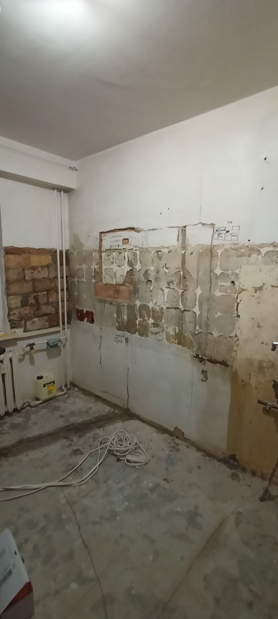 Pokój w trakcie demontażu starej okładziny ścian, widoczne fragmenty cegieł, instalacje i notatki na ścianie. Przygotowanie do prac wykończeniowych.