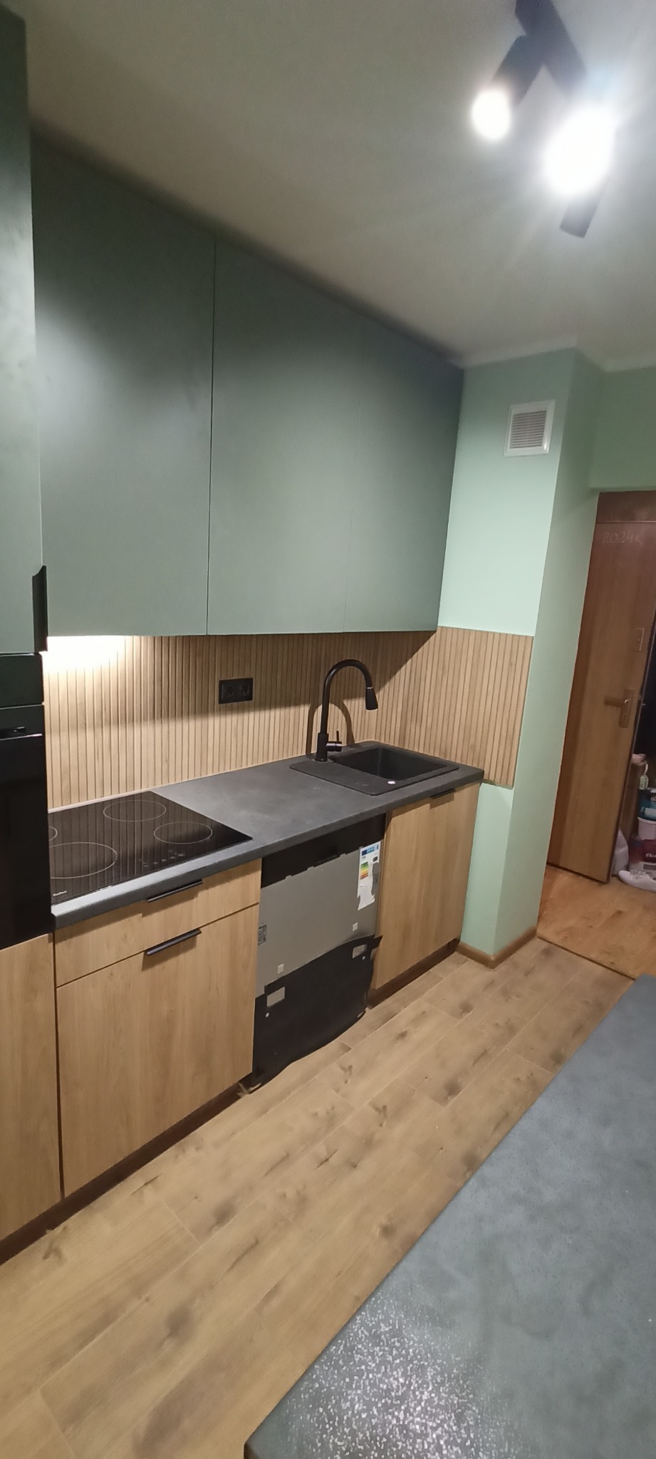 Nowoczesna kuchnia z zielonymi szafkami, drewnianymi frontami i blatem w odcieniach szarości. Uwagę zwraca drewniana okładzina ścienna i czarna armatura.