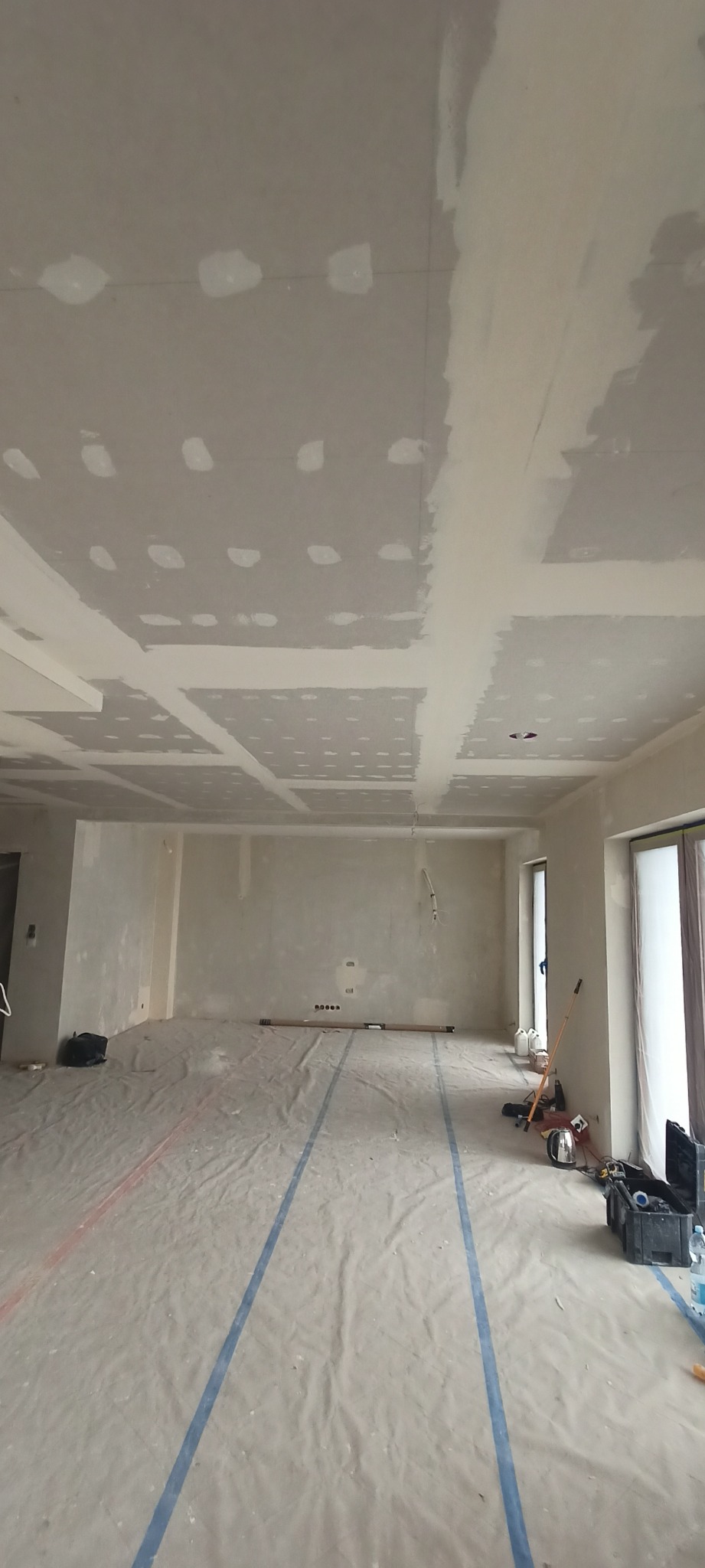 Wnętrze w trakcie prac wykończeniowych: sufit z płyt gipsowo-kartonowych, częściowo zaszpachlowany, podłoga zabezpieczona folią malarską, widoczne narzędzia.