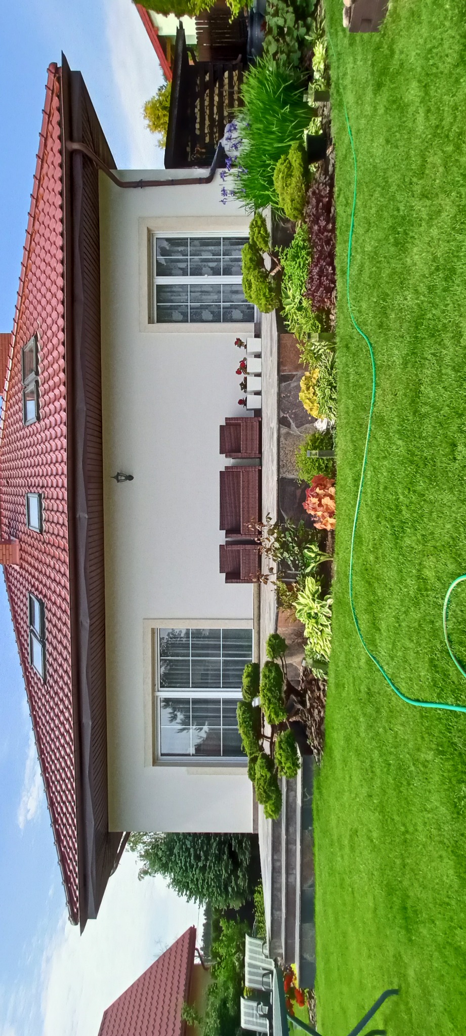 Elewacja domu z czerwonym dachem, widok na taras z meblami ogrodowymi i zadbany trawnik z rabatami kwiatowymi. Jasna fasada, okna z białymi ramami.