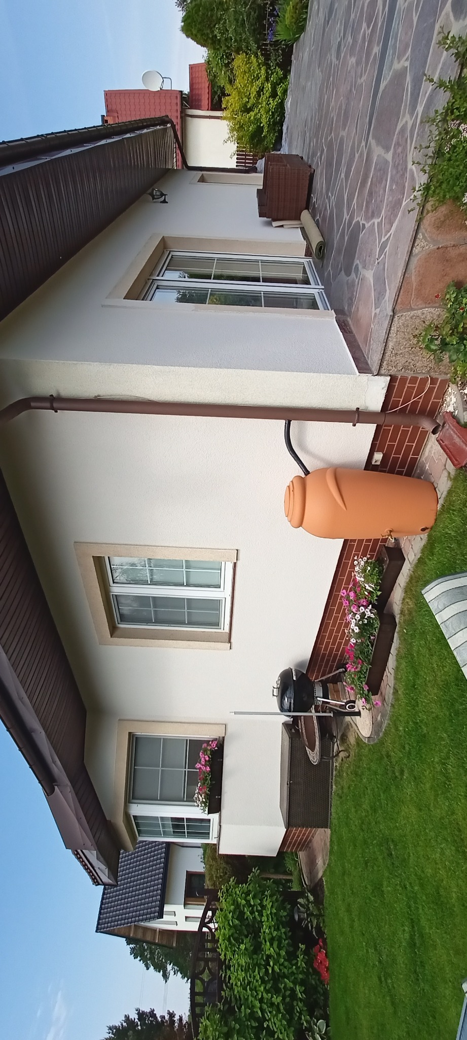 Elewacja domu po malowaniu z brązową rynną i zbiornikiem na deszczówkę. Widoczne okna z kwiatami i kamienny chodnik. Jasna kolorystyka, spokojne otoczenie.