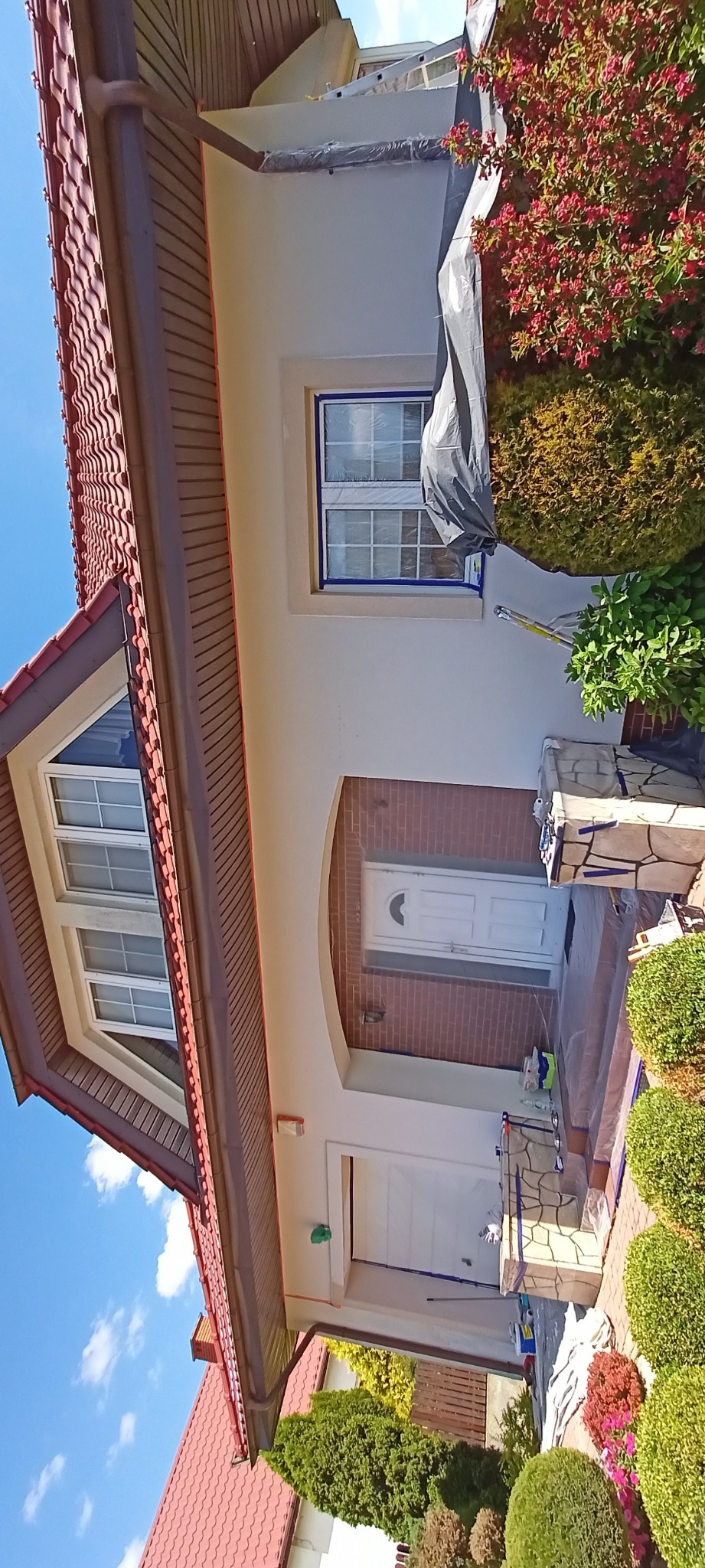 Elewacja domu w trakcie malowania: okna zabezpieczone folią, widoczne narzędzia malarskie, świeżo pomalowana ściana. Częściowo zakryte krzewy.
