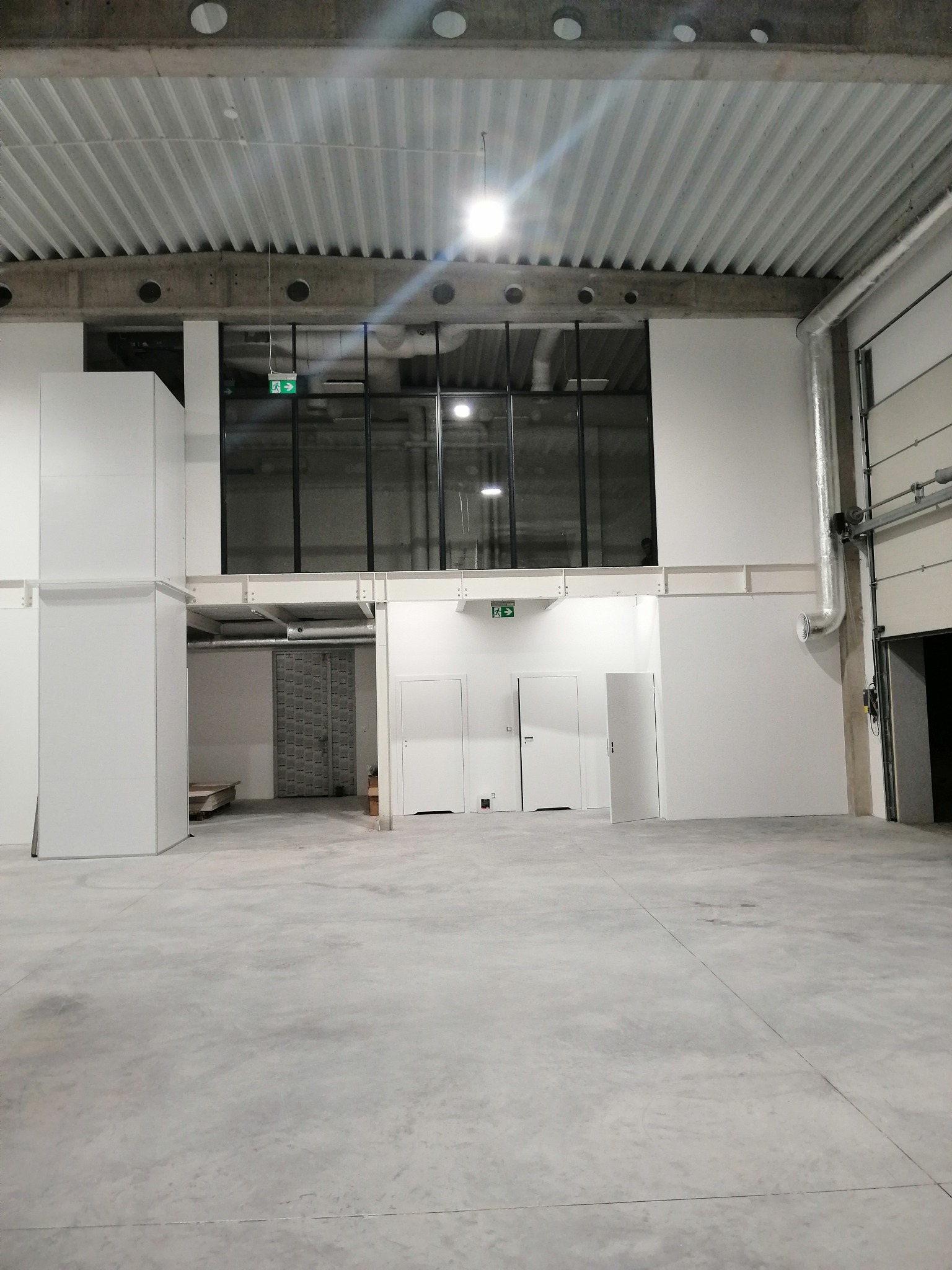 Surowe wnętrze biurowe w stanie deweloperskim z betonową podłogą, białymi ścianami, widoczną instalacją wentylacyjną, oknami i drzwiami, oświetlone górnym światłem.