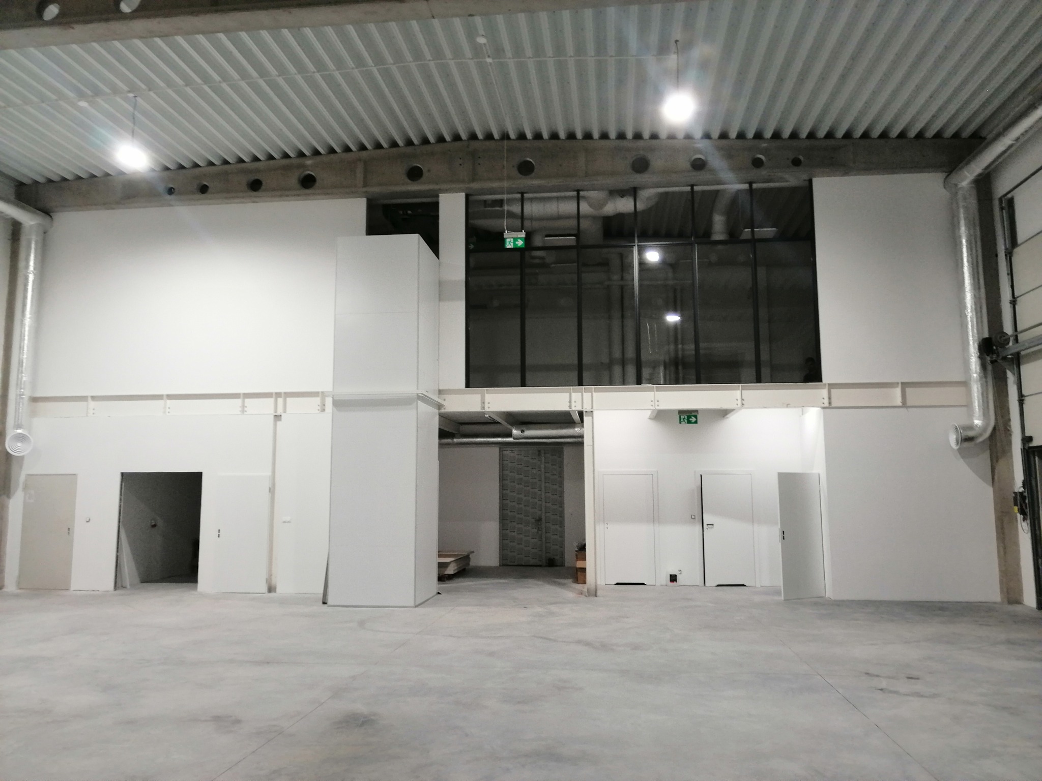 Przestronne, puste wnętrze hali produkcyjnej lub magazynowej z białymi ścianami, kilkoma białymi drzwiami, betonową podłogą i widoczną instalacją pod sufitem. Okno z czarnymi ramami w górnej części...