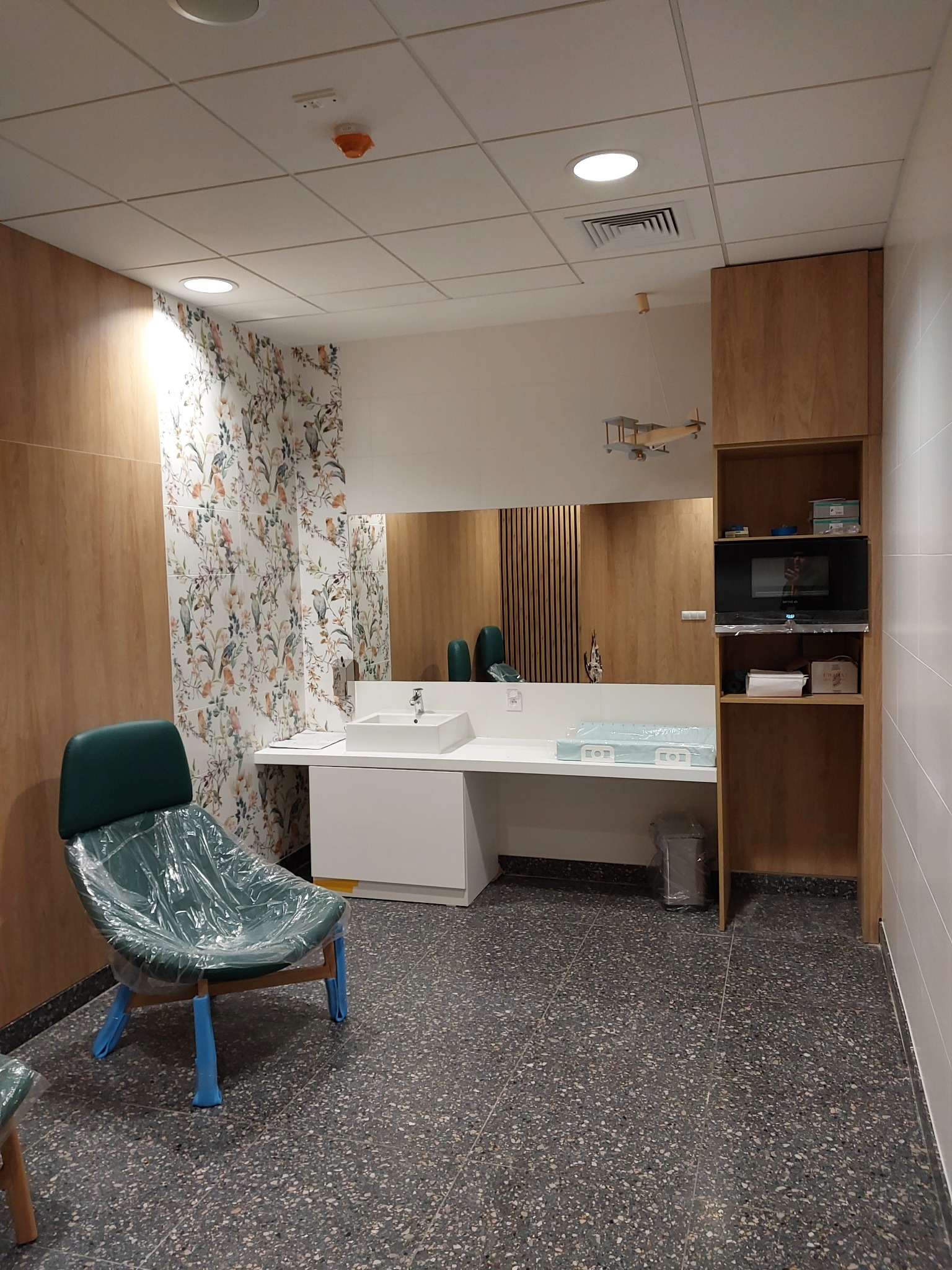 Nowoczesna łazienka w biurze z drewnianymi panelami, wzorzystą tapetą, białym blatem z umywalką, zielonym krzesłem zabezpieczonym folią i ozdobnym modelem samolotu.