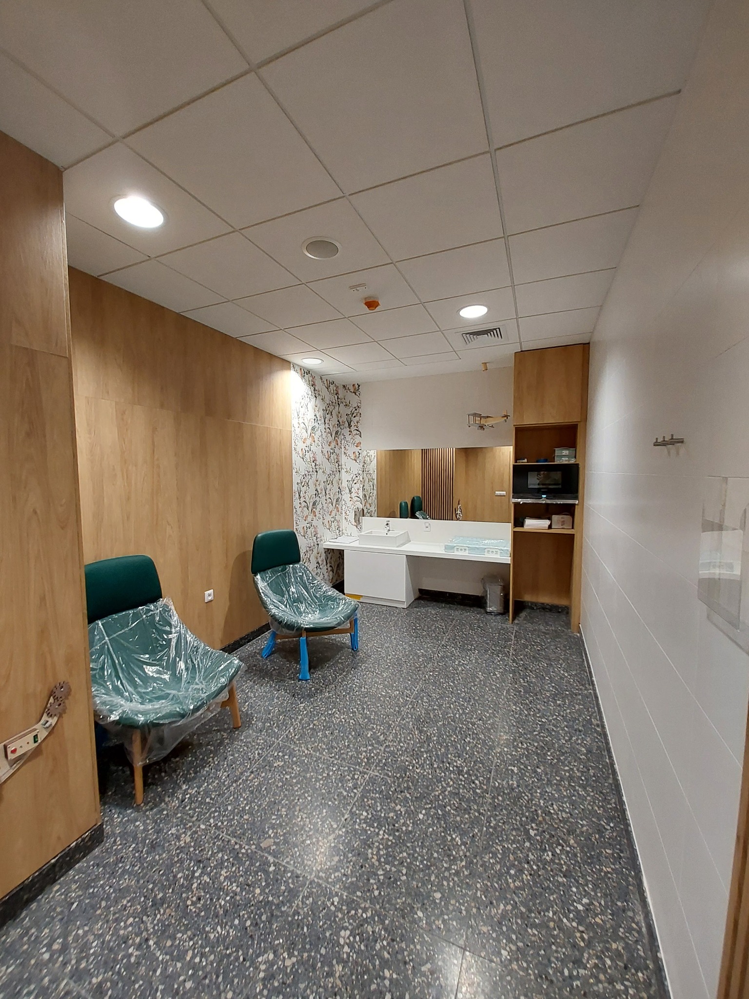 Nowoczesna poczekalnia biurowa z drewnianą okładziną ścienną, dwoma krzesłami zabezpieczonymi folią ochronną, umywalkami i szafką z mikrofalówką, wykończona szarą posadzką lastryko.