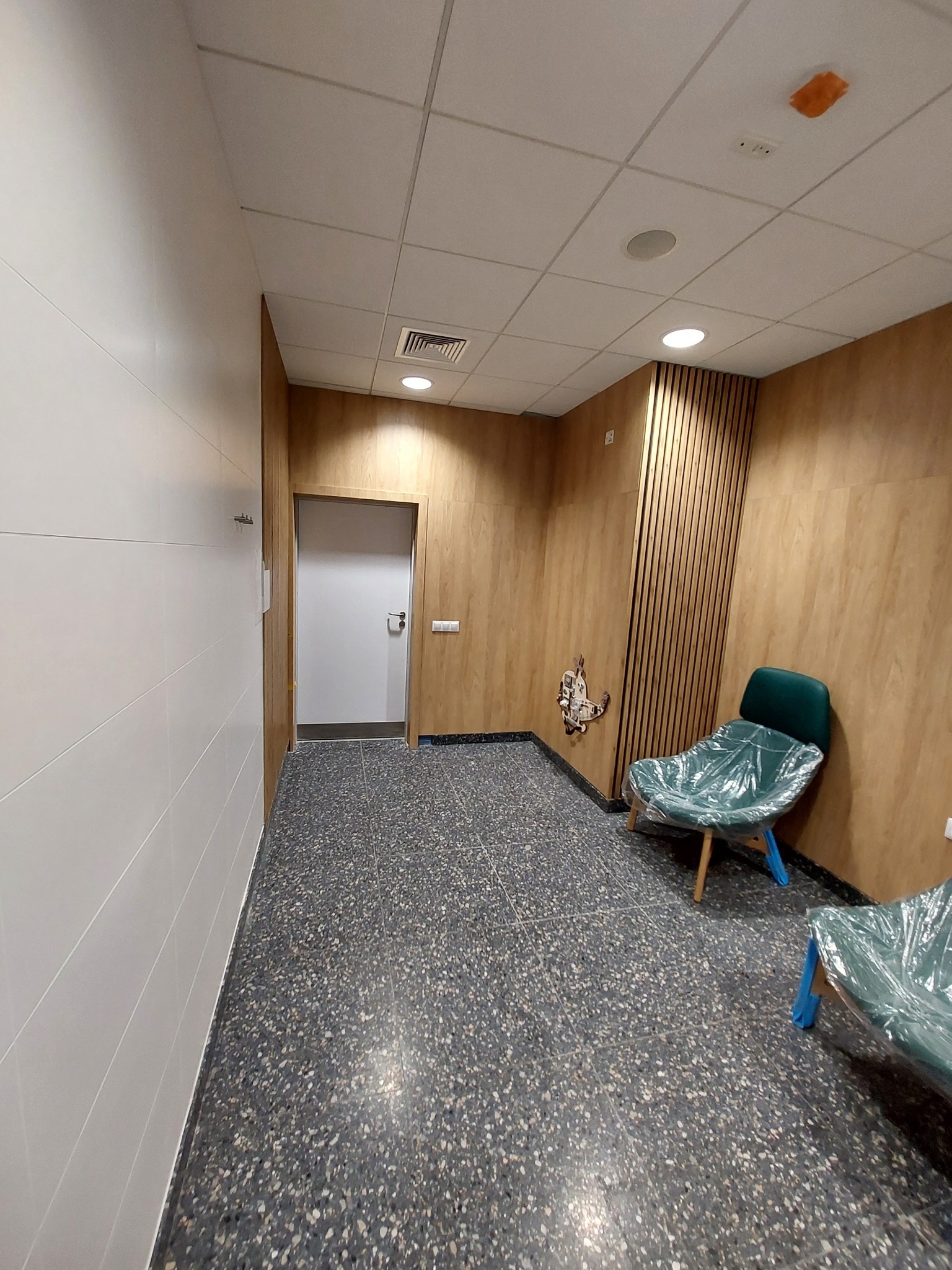 Korytarz biurowy po remoncie z wykończonymi ścianami w panelach drewnopodobnych, nową podłogą z lastryko i zabezpieczonymi folią krzesłami.