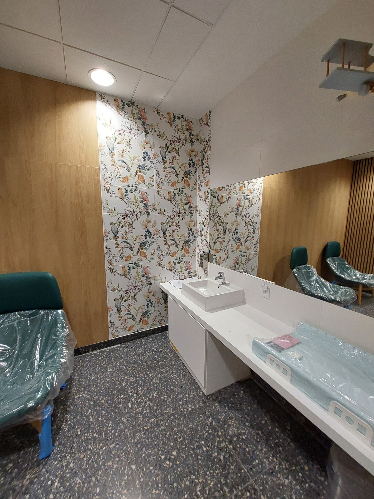 Jasne wnętrze z dekoracyjną tapetą w kwiaty i ptaki, białą umywalką i blatem, oraz krzesłami zabezpieczonymi folią, prawdopodobnie w trakcie lub po remoncie.