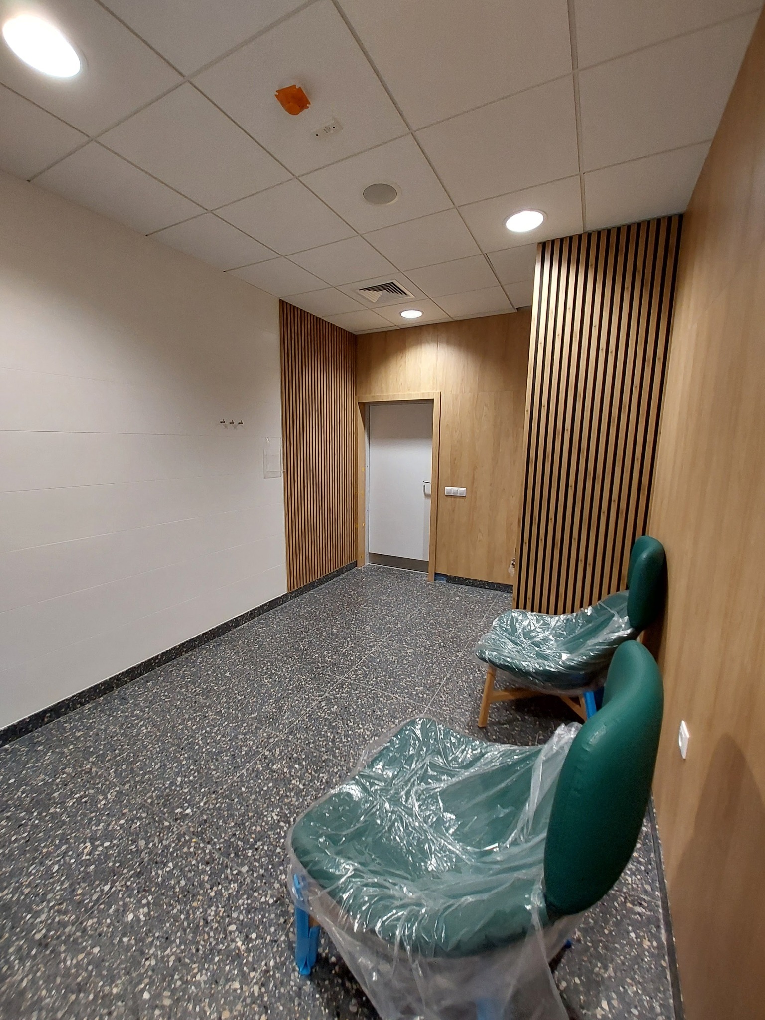 Korytarz biurowy z drewnianymi panelami ściennymi, szarą podłogą z lastryko i dwoma krzesłami zabezpieczonymi folią ochronną przed zabrudzeniami podczas prac wykończeniowych.