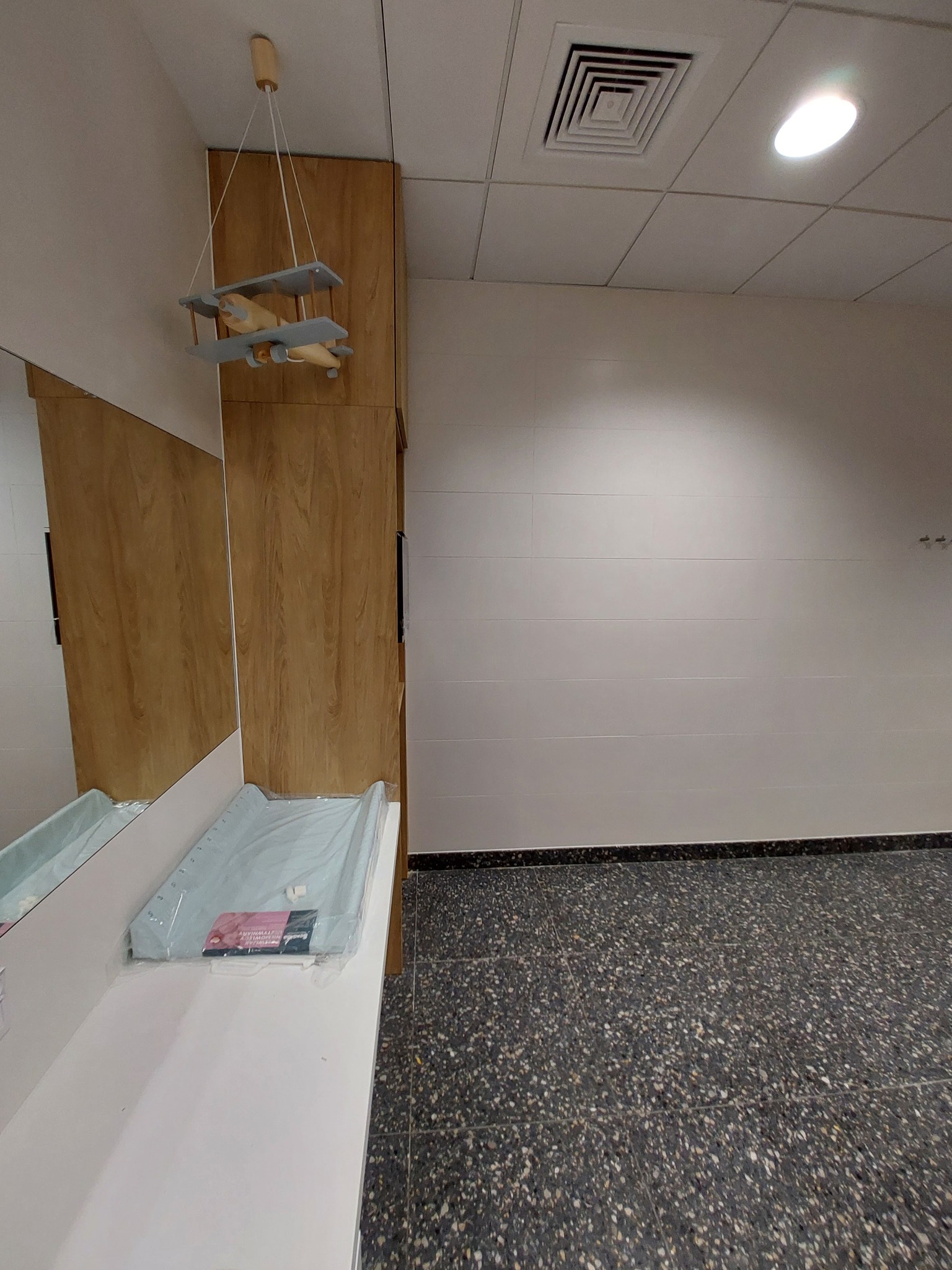 Wykończona łazienka biurowa z drewnianą zabudową szafki umywalkowej, białą umywalką z przewijakiem i wiszącą dekoracją w kształcie samolotu.