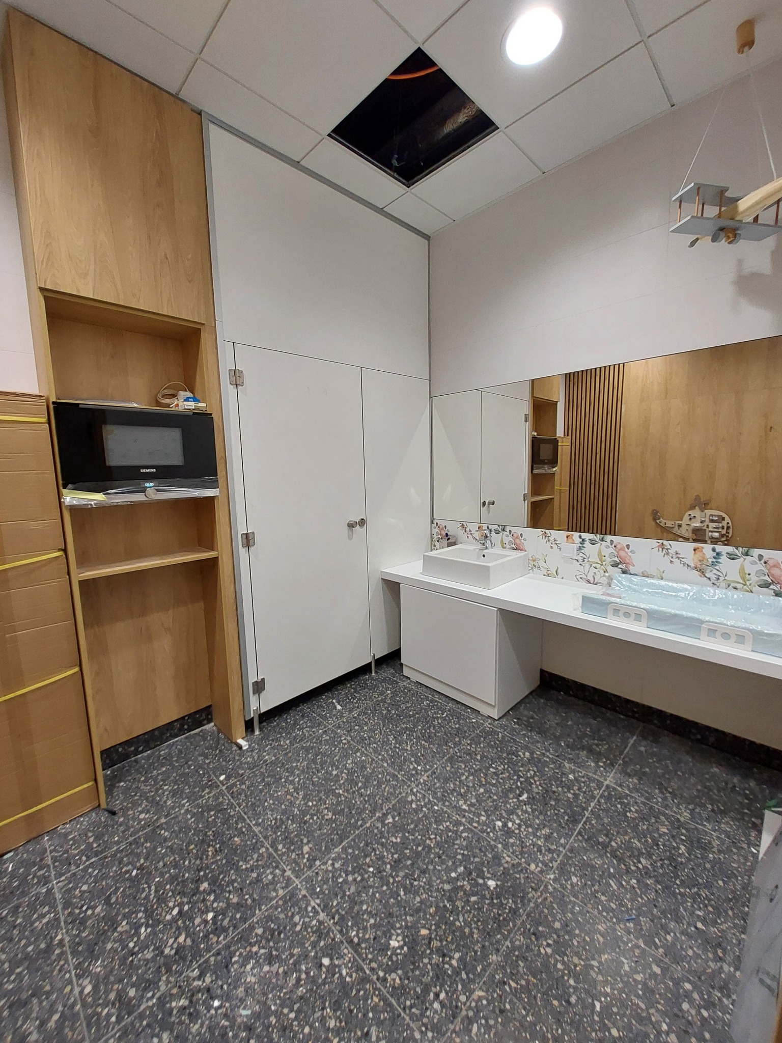 Wykończona łazienka biurowa z drewnianymi akcentami, białą armaturą i szafkami, oraz dekoracyjnymi płytkami z motywem ptaków nad umywalką. Widoczny zamontowany piekarnik w drewnianej zabudowie...