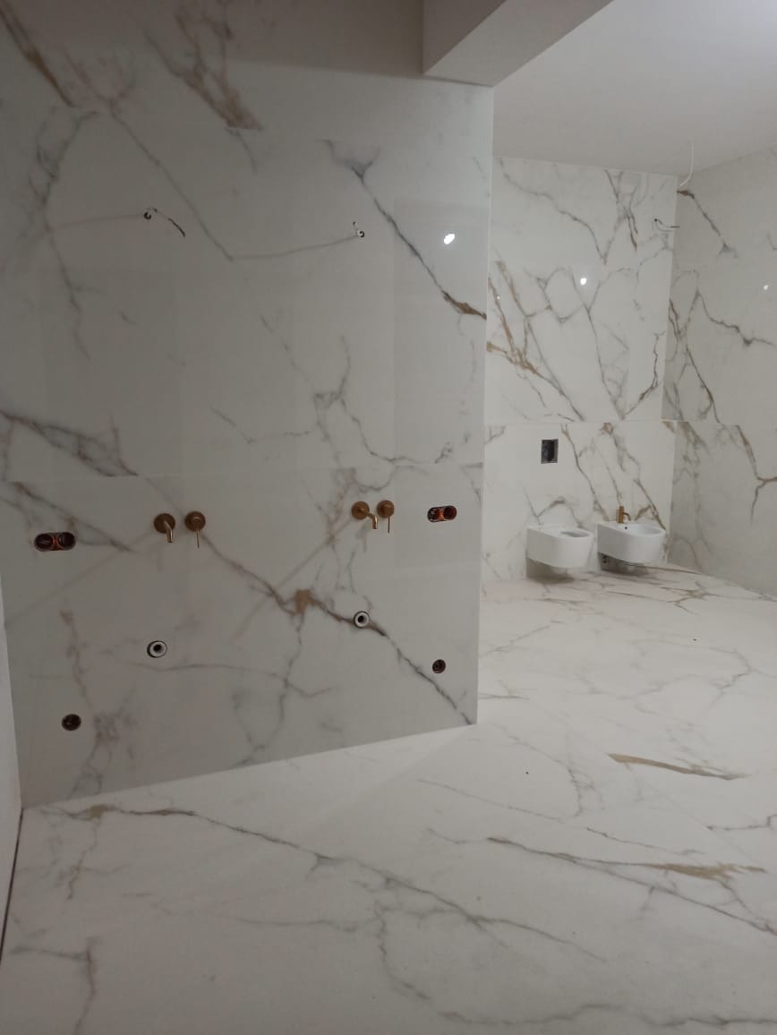 Wykończona łazienka z białymi płytkami imitującymi marmur na ścianach i podłodze, widoczne przygotowane punkty instalacji wodnej i dwa białe bidety.