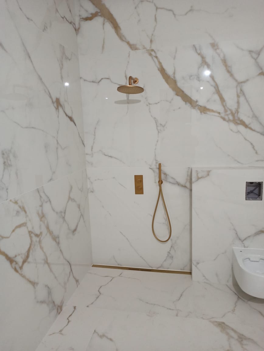 Kabina prysznicowa wyłożona białymi płytkami imitującymi marmur z złotymi żyłami, z zamontowaną złotą armaturą i odpływem liniowym w kolorze złota.