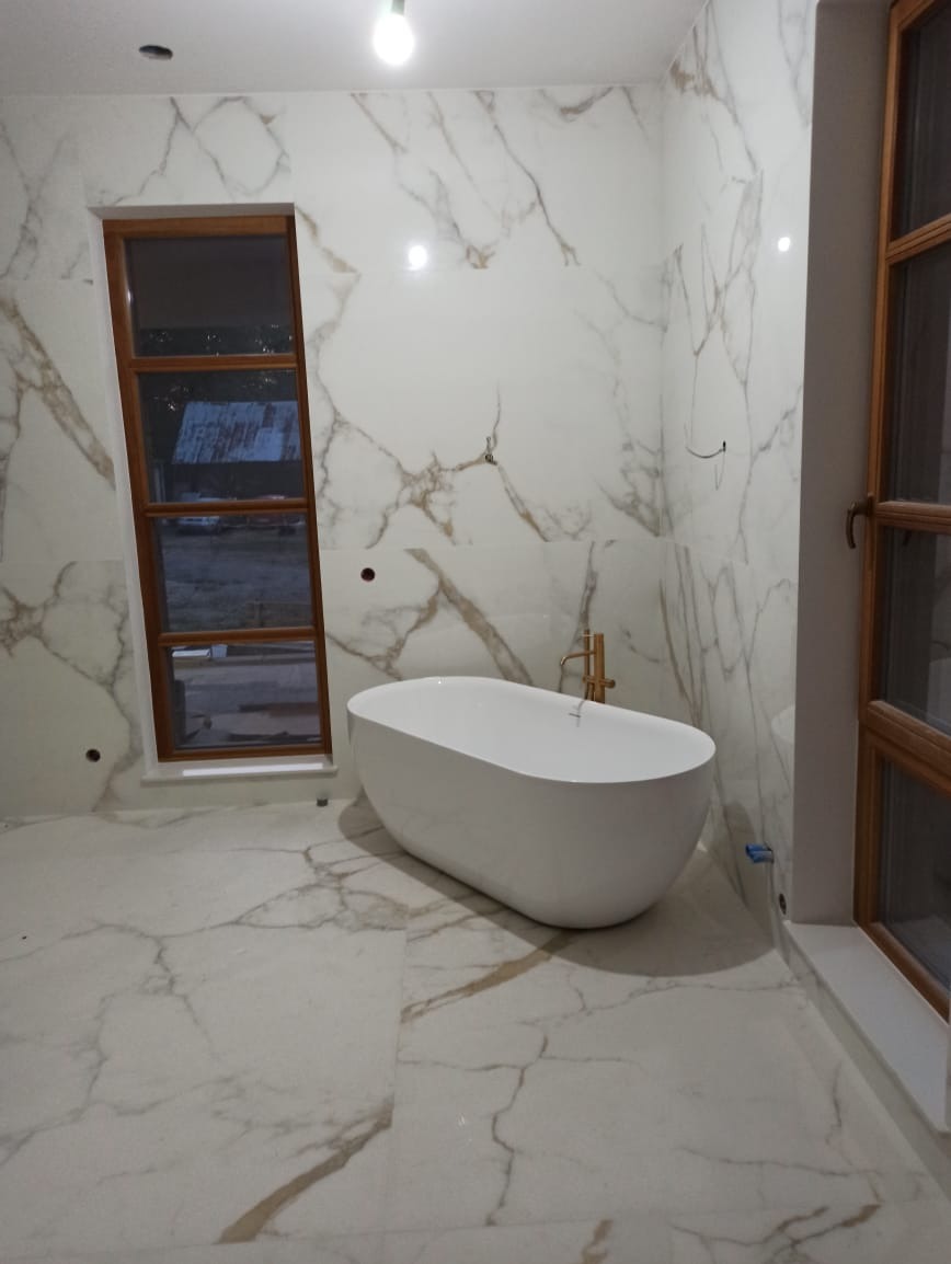 Nowoczesna łazienka z wolnostojącą białą wanną na tle marmurowych płytek, z drewnianą ramą okienną i złotą armaturą.