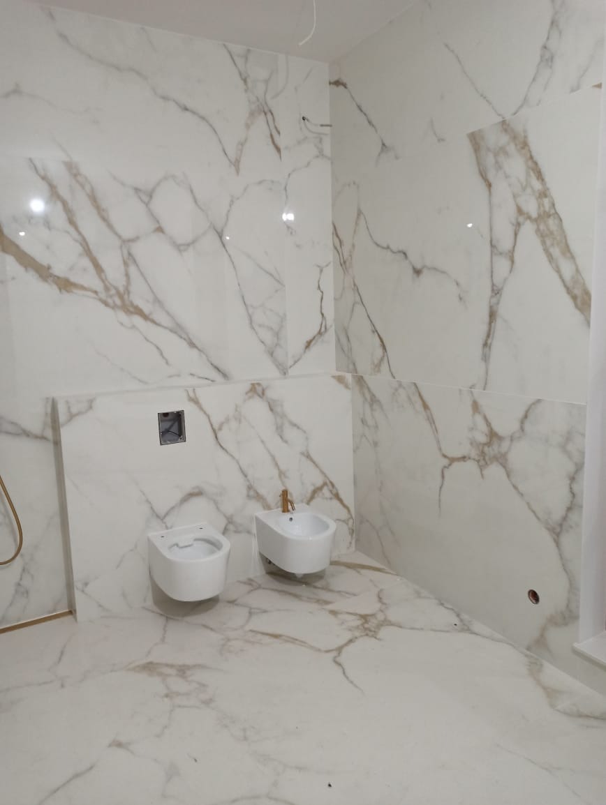 Łazienka z białymi płytkami imitującymi marmur z brązowymi żyłami, wisząca toaleta i bidet z chromowaną baterią, widoczny otwór w ścianie pod instalację.