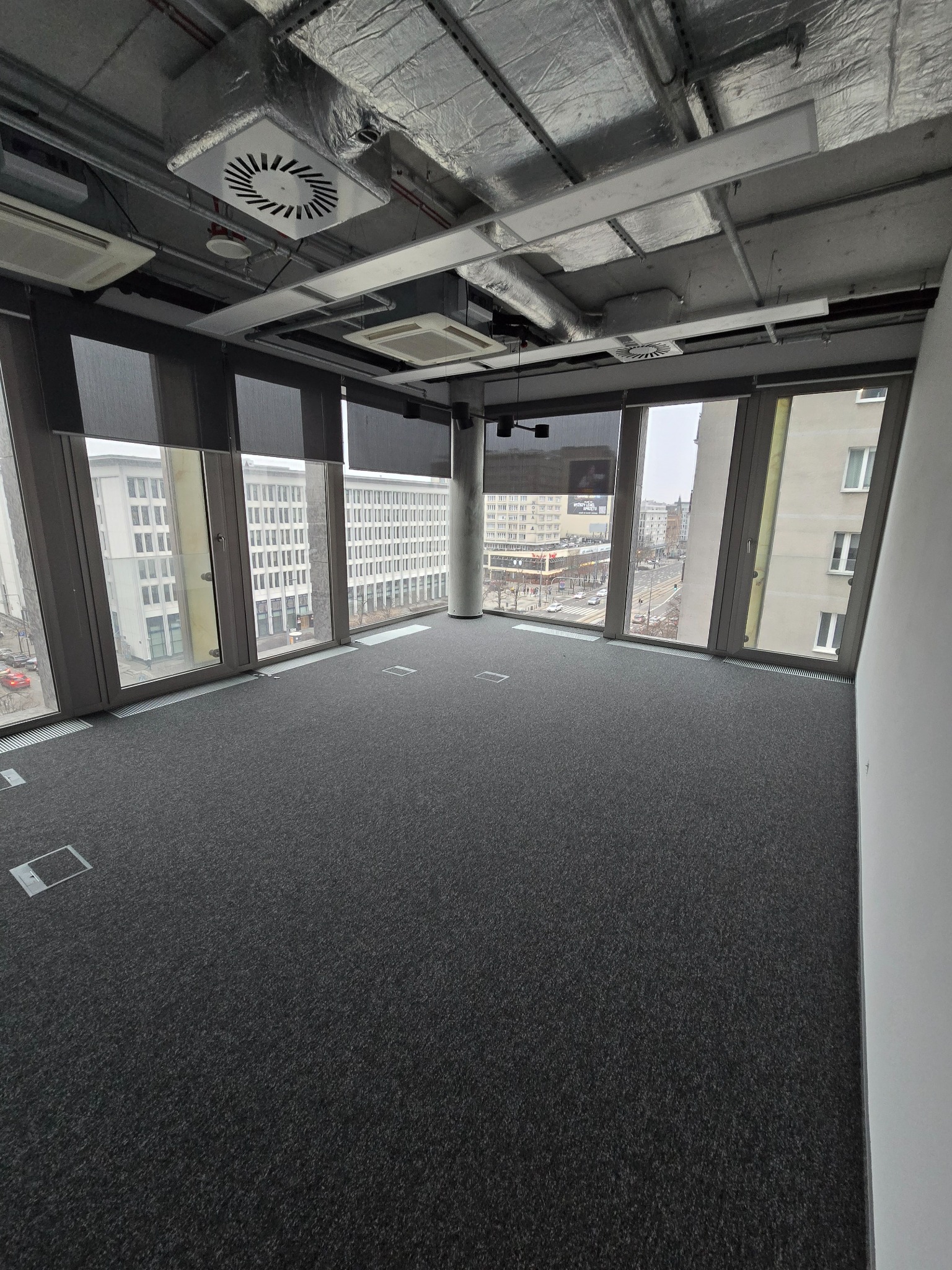 Puste, odnowione biuro z szarą wykładziną, widokiem na miasto przez duże okna z roletami, i odsłoniętym sufitem z instalacjami wentylacyjnymi.