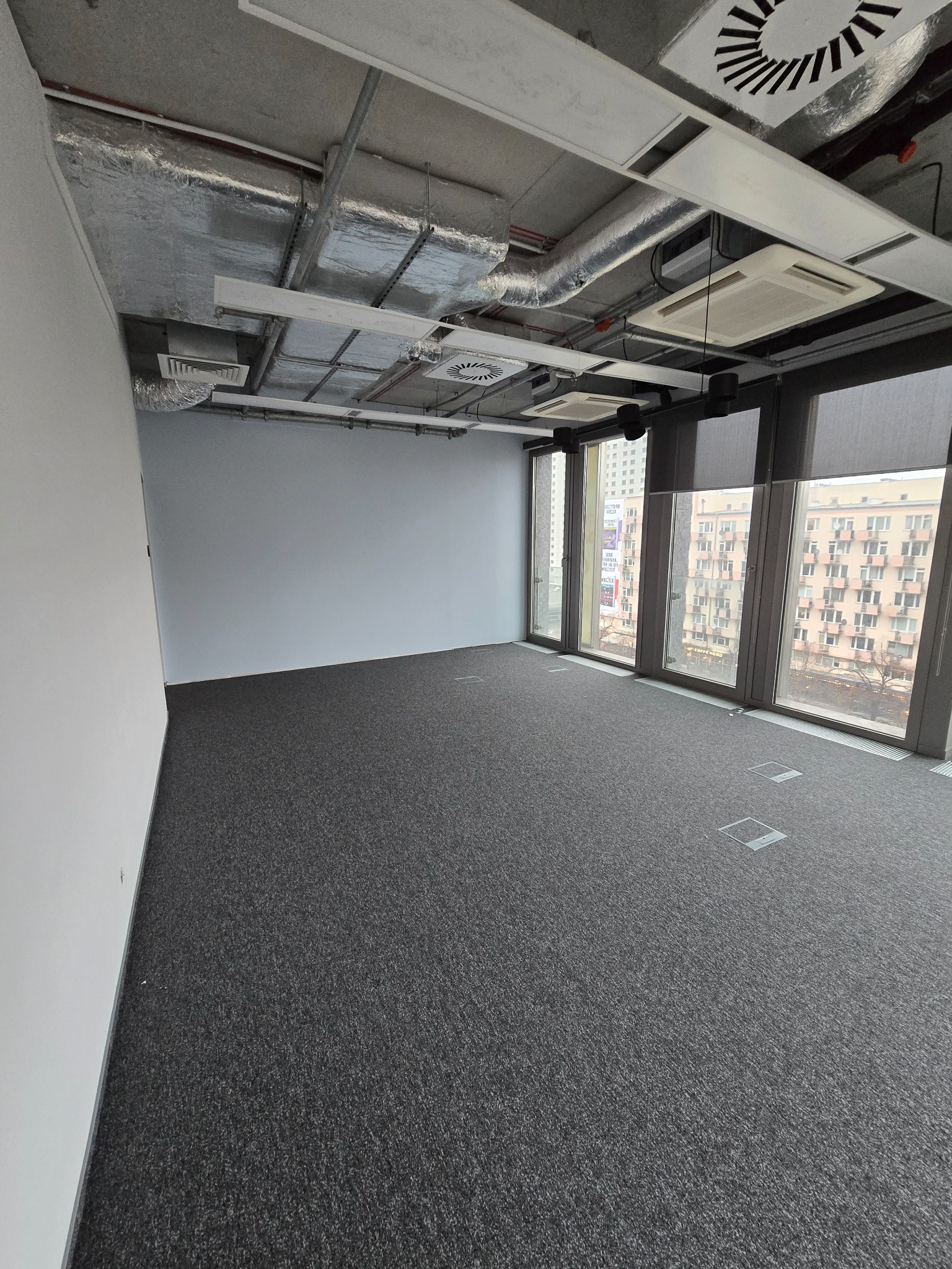 Puste biuro z szarą wykładziną, widoczne okna z roletami, sufit z instalacjami i klimatyzacją, ściany pomalowane na jasny kolor.