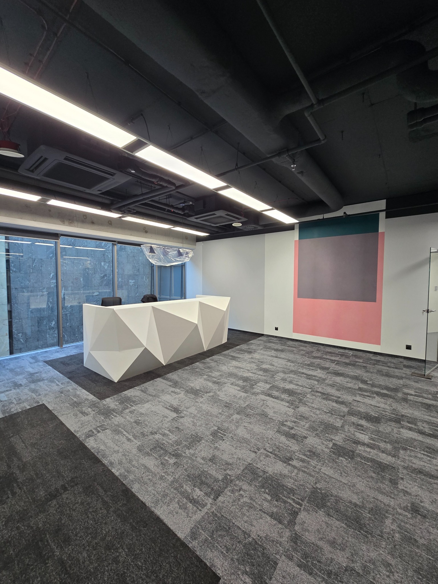 Nowoczesne biuro z geometryczną recepcją, wzorzystą wykładziną dywanową i minimalistycznym malowaniem ścian w kolorach różu, szarości i zieleni.