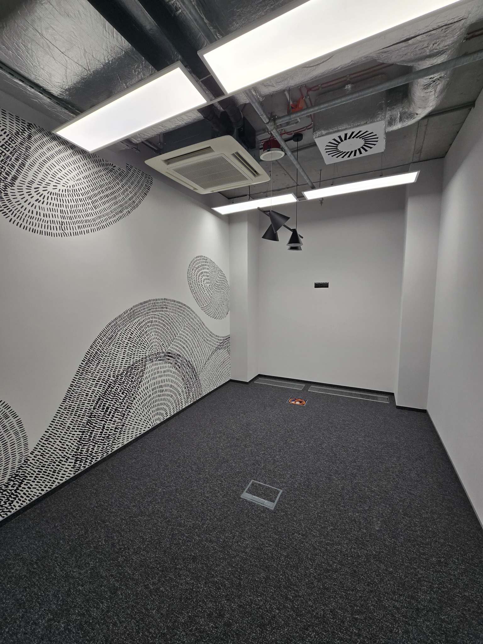 Puste biuro z szarą wykładziną, białe ściany z abstrakcyjną grafiką w stylu dymków, sufit z widocznymi instalacjami i nowoczesnym oświetleniem LED oraz klimatyzacją, oświetlenie punktowe.