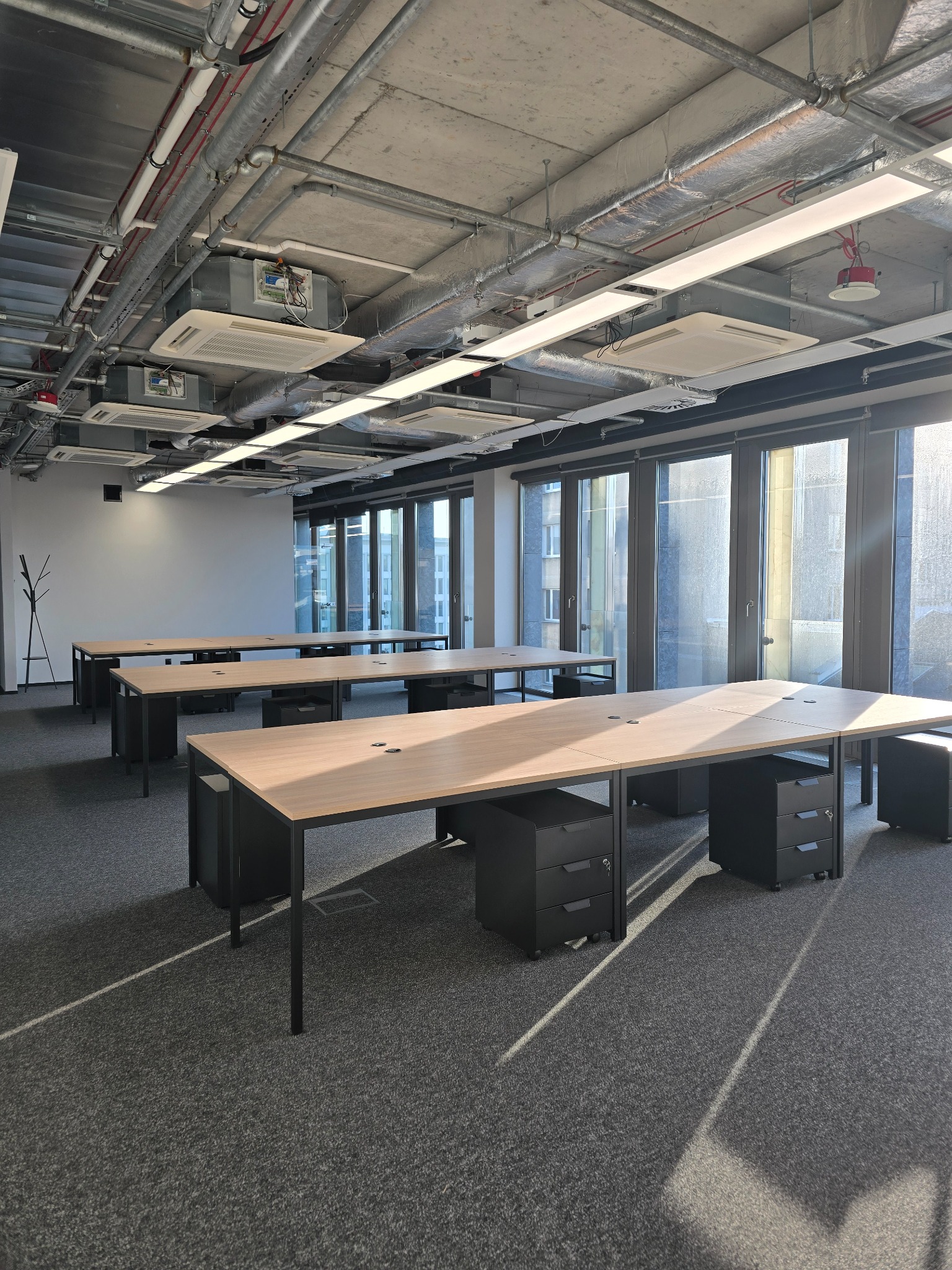 Jasne biuro po remoncie z rzędami biurek z czarnymi kontenerkami, widoczne instalacje pod sufitem i duże okna.