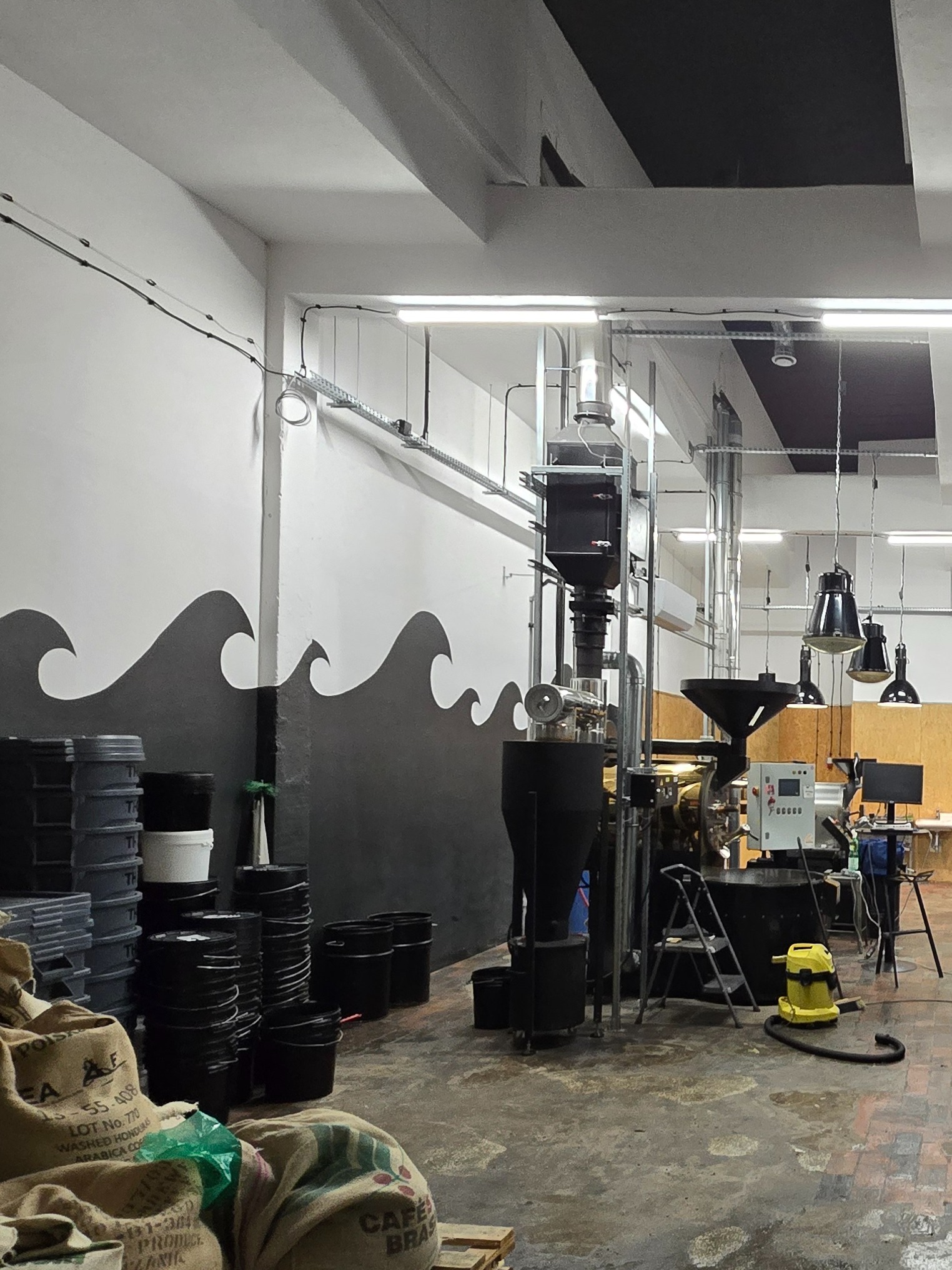 Wnętrze przemysłowe z maszyną do palenia kawy, workami z ziarnami i pomalowaną ścianą w fale. Widoczne elementy instalacji elektrycznej i oświetlenie.