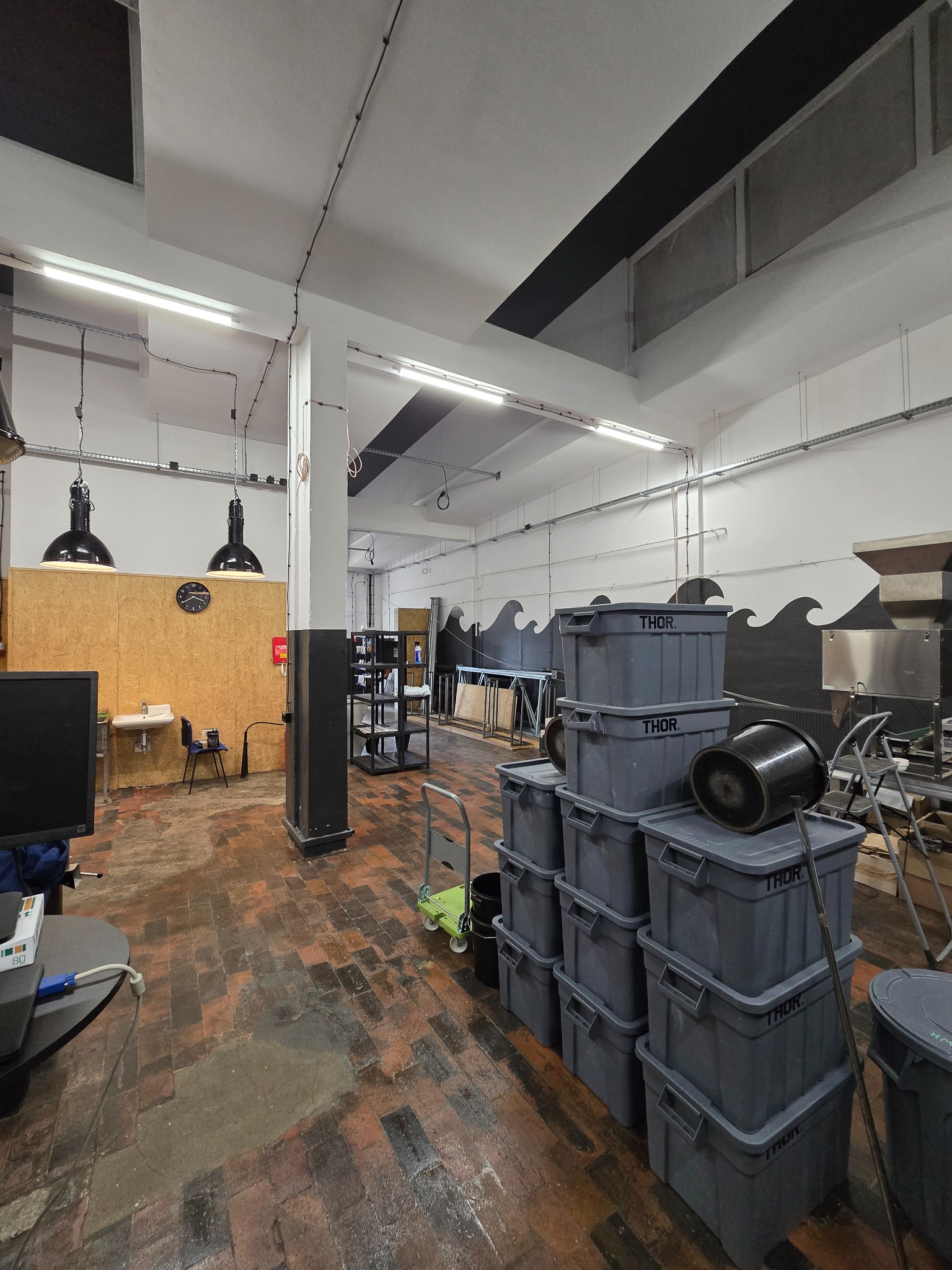 Przestronne wnętrze biura z ceglaną podłogą, wyposażone w lampy przemysłowe i szare pojemniki THOR. Ściany zdobi motyw fal. Widoczne elementy wyposażenia biurowego.