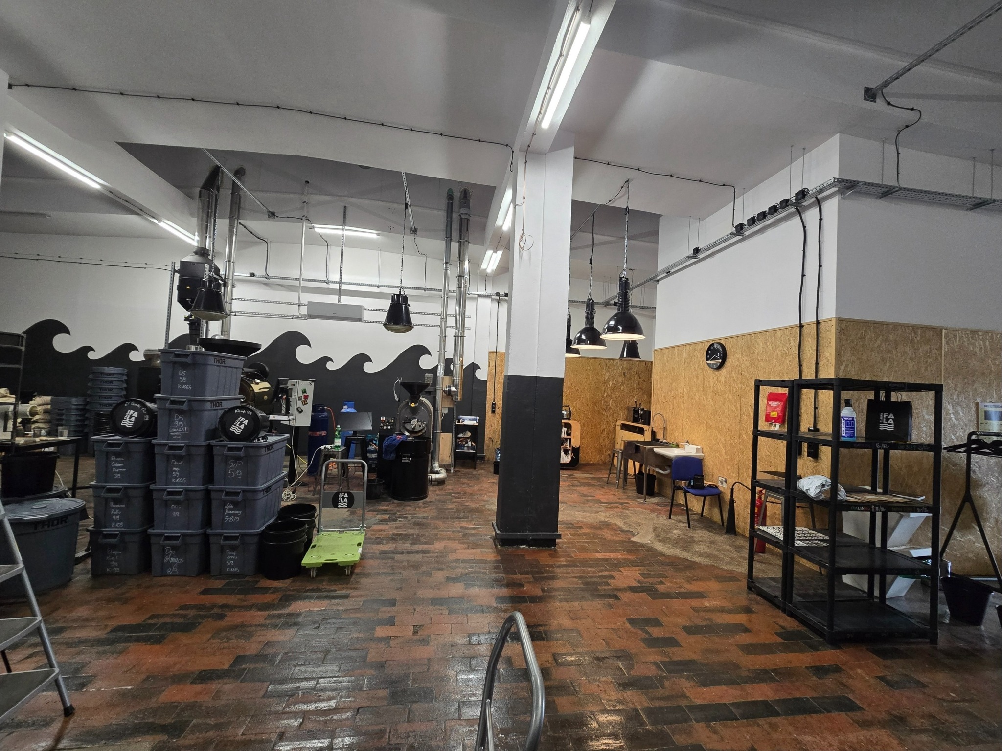 Przestronne wnętrze biurowe z widoczną instalacją elektryczną pod sufitem, lampami i elementami dekoracyjnymi w postaci fal na ścianie. Widoczne regały i wyposażenie.