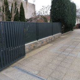 Ogro-Zach Ogrodzenia - Nowoczesne, grafitowe ogrodzenie panelowe z otwartą furtką, osadzone na niskim murku z bloczków imitujących kamień, na tle szarej kostki brukowej i zieleni.