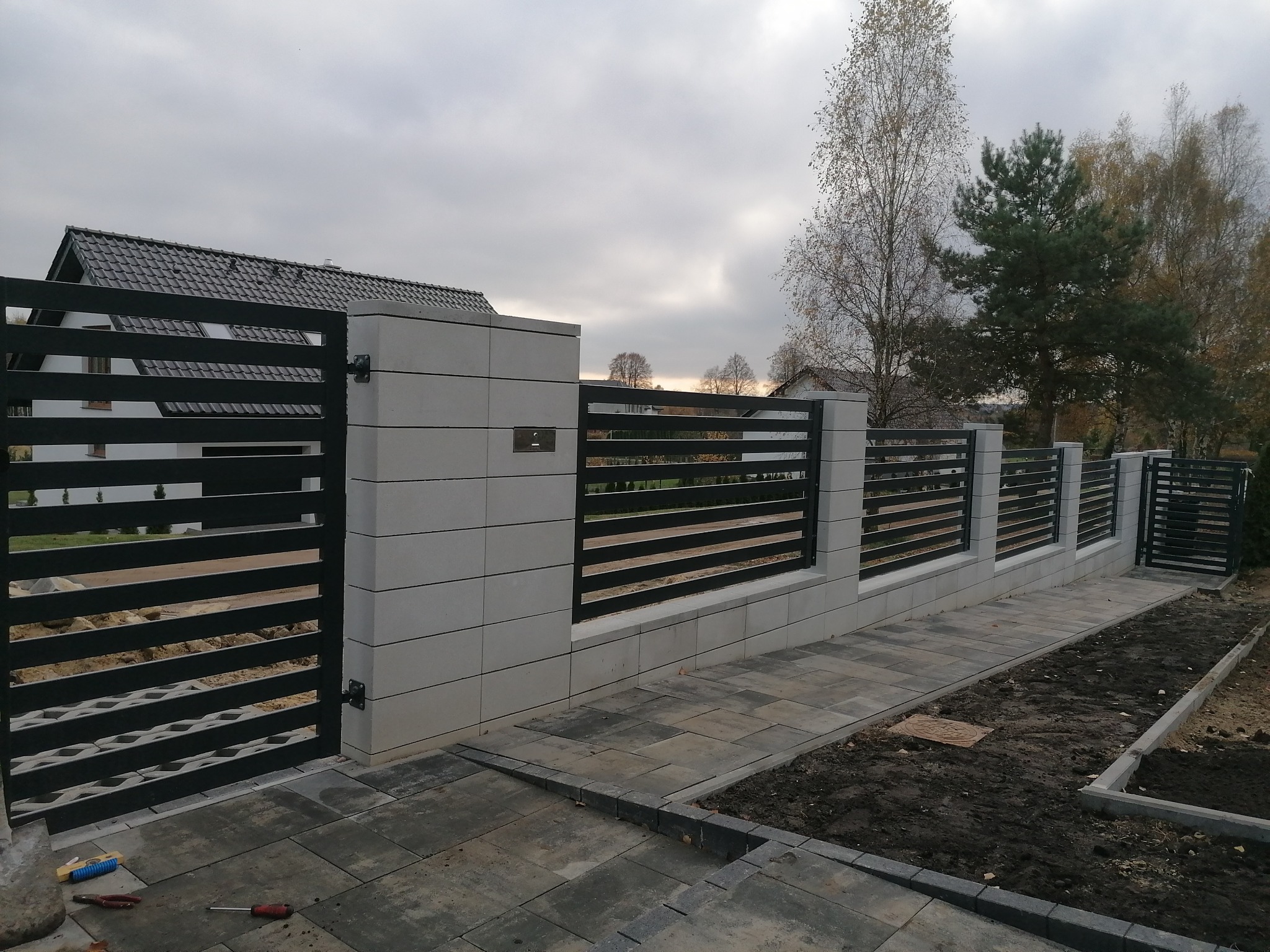 Nowoczesne ogrodzenie panelowe z bramą przesuwną i słupkami z szarej kostki betonowej, widok z zewnątrz posesji, na ziemi leżą narzędzia.