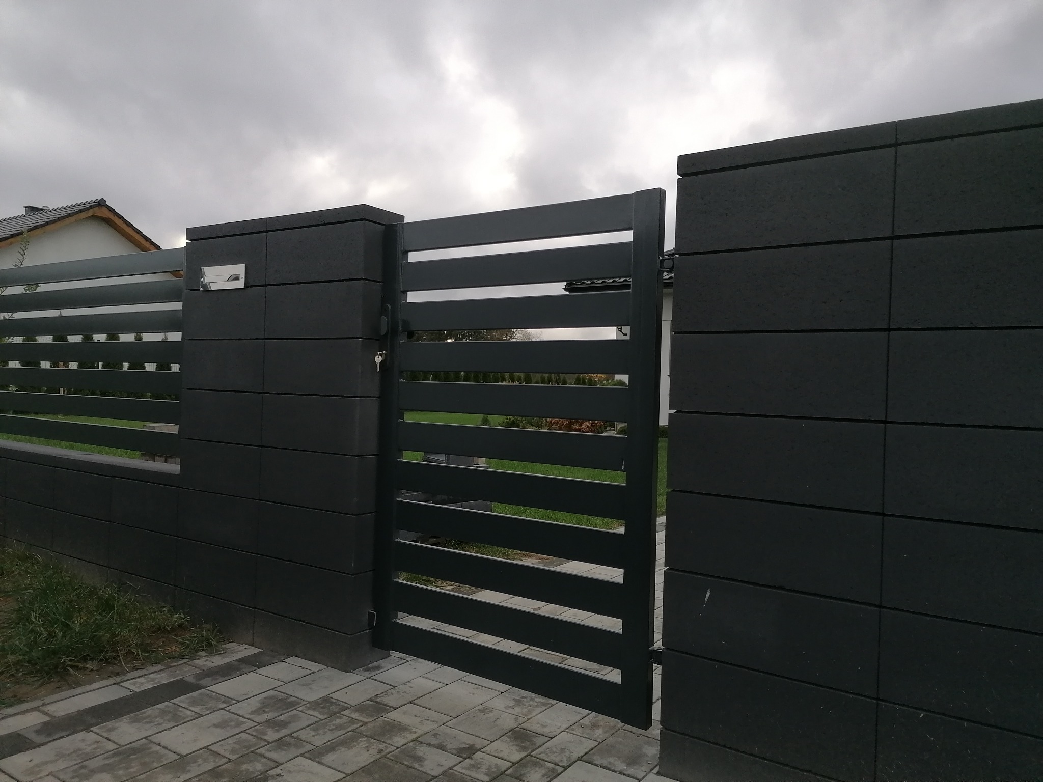 Nowoczesna, antracytowa brama przesuwna z poziomymi szczeblami, wbudowana w ogrodzenie z betonowych bloczków, skrzynka na listy wbudowana w słupek bramy, kostka brukowa na podjeździe, pochmurne...