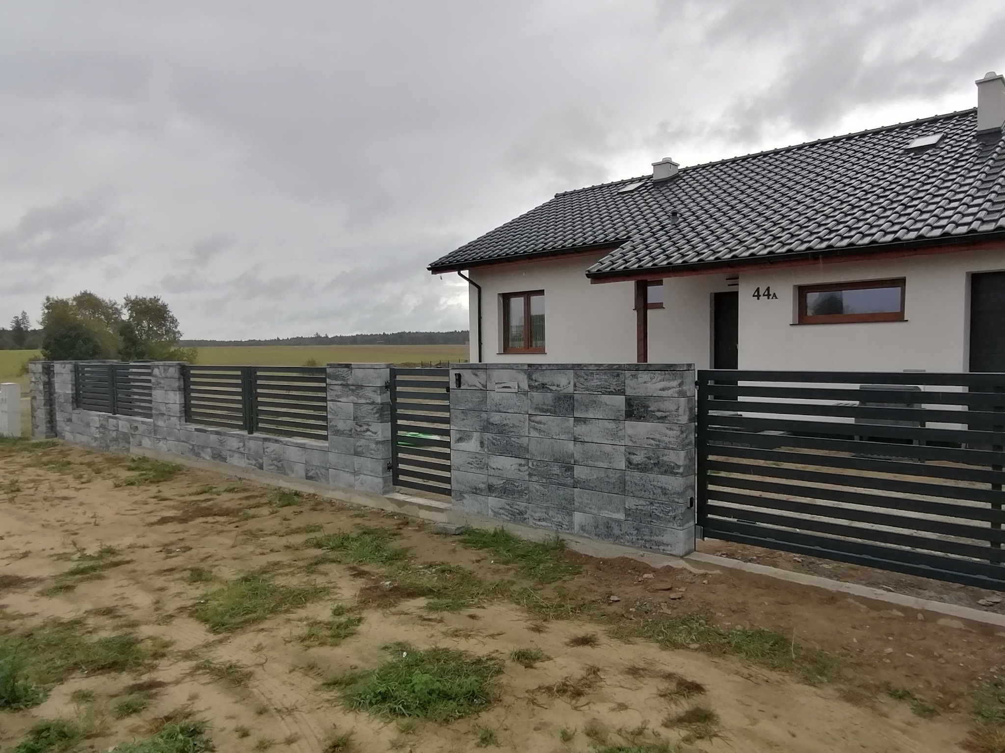Nowoczesne ogrodzenie panelowe z bramą przesuwną, wykończone szarymi bloczkami betonowymi, z widokiem na dom z ciemną dachówką i polami w tle, pochmurny dzień.