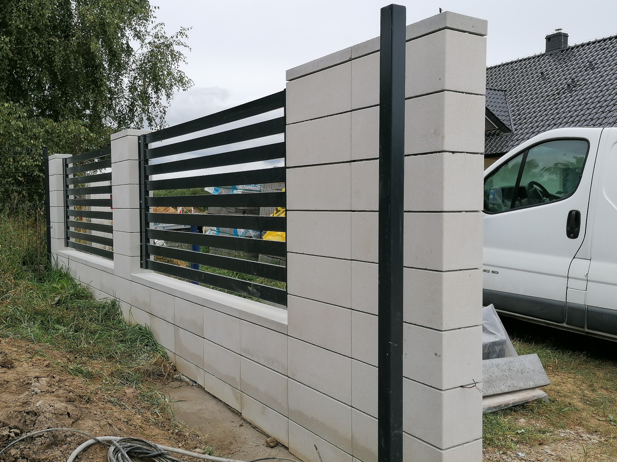 Nowoczesne ogrodzenie z szarego betonu architektonicznego z poziomymi, czarnymi przęsłami, widoczny fragment białego samochodu dostawczego i niezagospodarowany teren.
