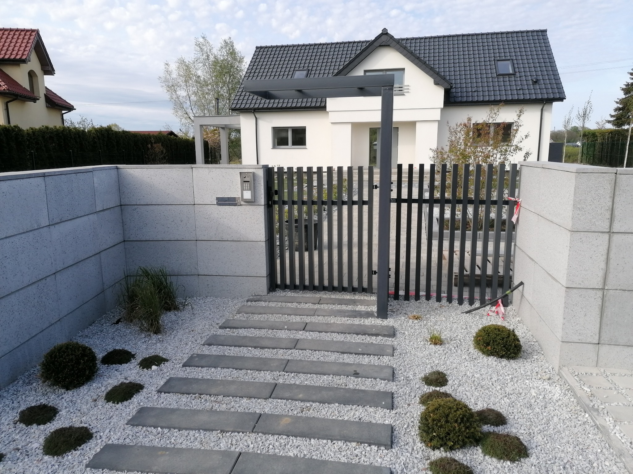 Nowoczesna, grafitowa brama furtka z domofonem przy wejściu do posesji w Gryficach, z betonowym ogrodzeniem i ścieżką z płyt na białym żwirze, prowadząca do domu z ciemnym dachem.