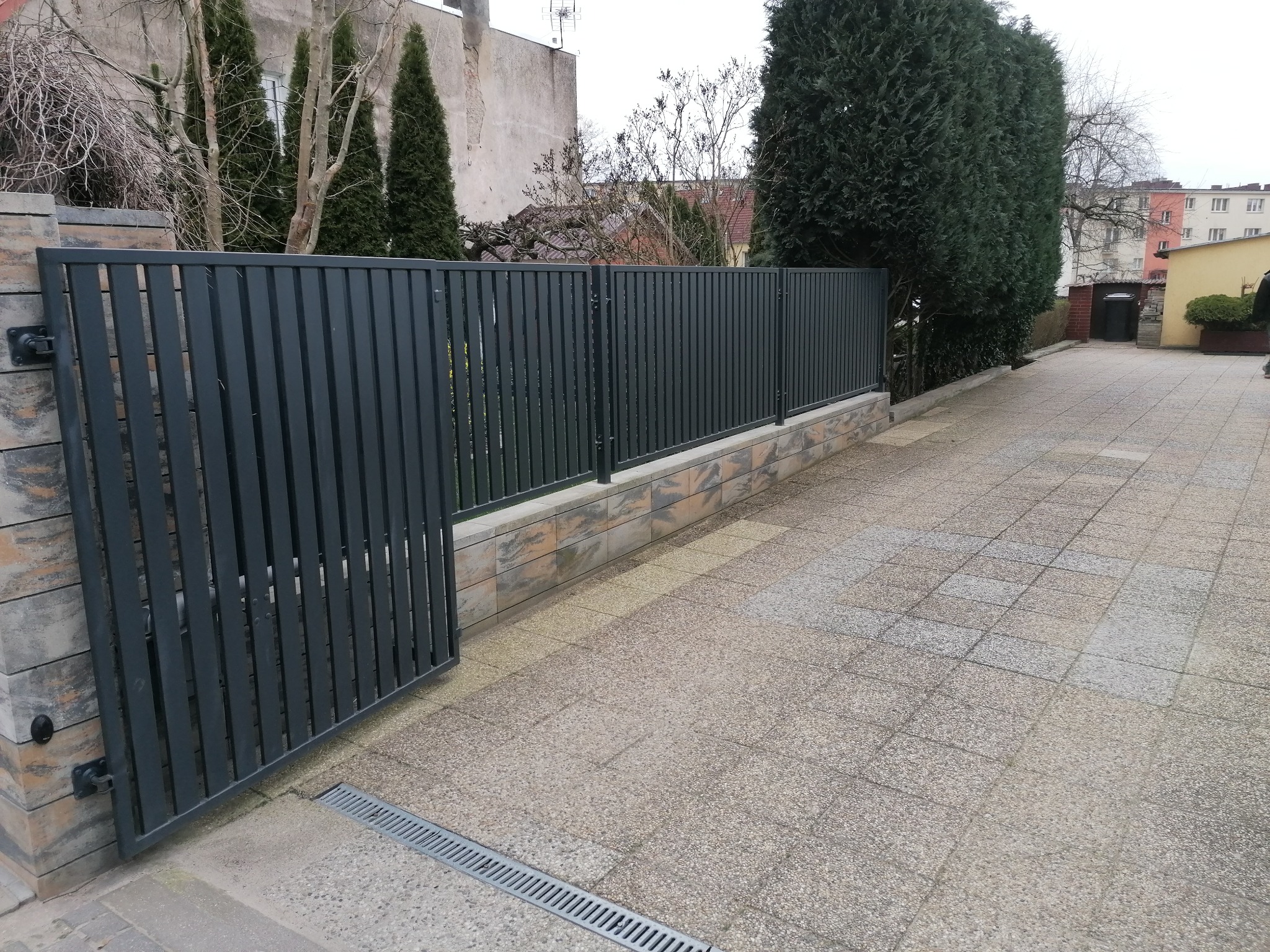 Nowoczesne, grafitowe ogrodzenie panelowe z otwartą furtką, osadzone na niskim murku z bloczków imitujących kamień, na tle szarej kostki brukowej i zieleni.