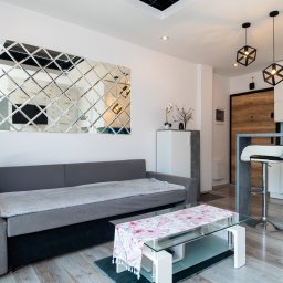 Bonus Nieruchomości - Nowoczesny salon z szarą sofą, lustrzaną ścianą i szklanym stolikiem kawowym. Oświetlenie stanowią geometryczne lampy wiszące. Widoczna kuchnia z barkiem i krzesłem.