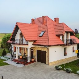 Bonus Nieruchomości - Dwupiętrowy dom z czerwonym dachem, balkonami z kutymi balustradami i garażem. Uwagę zwraca zadbany ogród z dekoracyjnymi krzewami i kostką brukową.